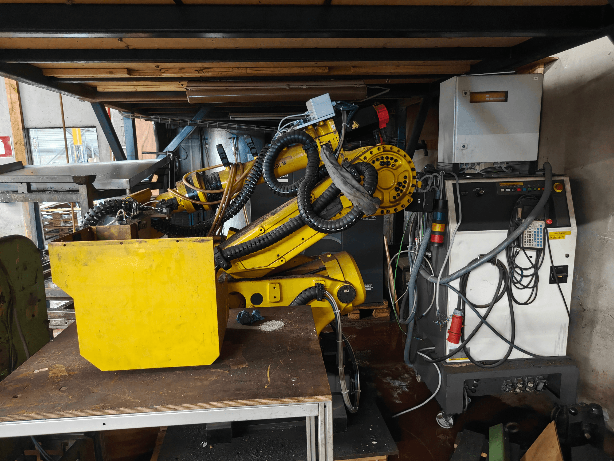 Widok z przodu ramienia robota FANUC S-430i W, w kolorze żółtym, wraz z panelem sterowania, kablami oraz żółtym elementem na stole warsztatowym.