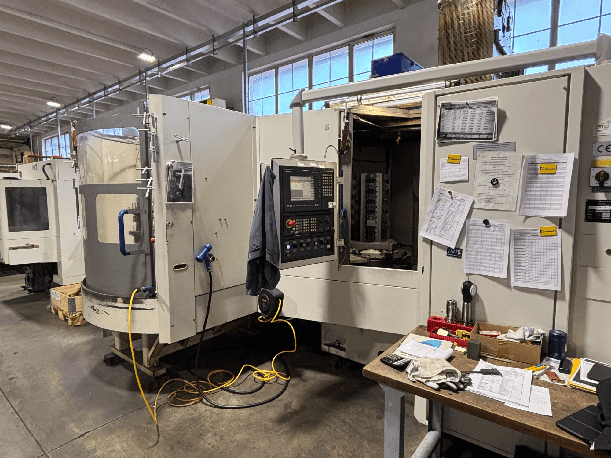 Maszyna CNC w warsztacie, widok z przodu, z panelem sterowania i dokumentacją z boku; narzędzia i materiały na stole.