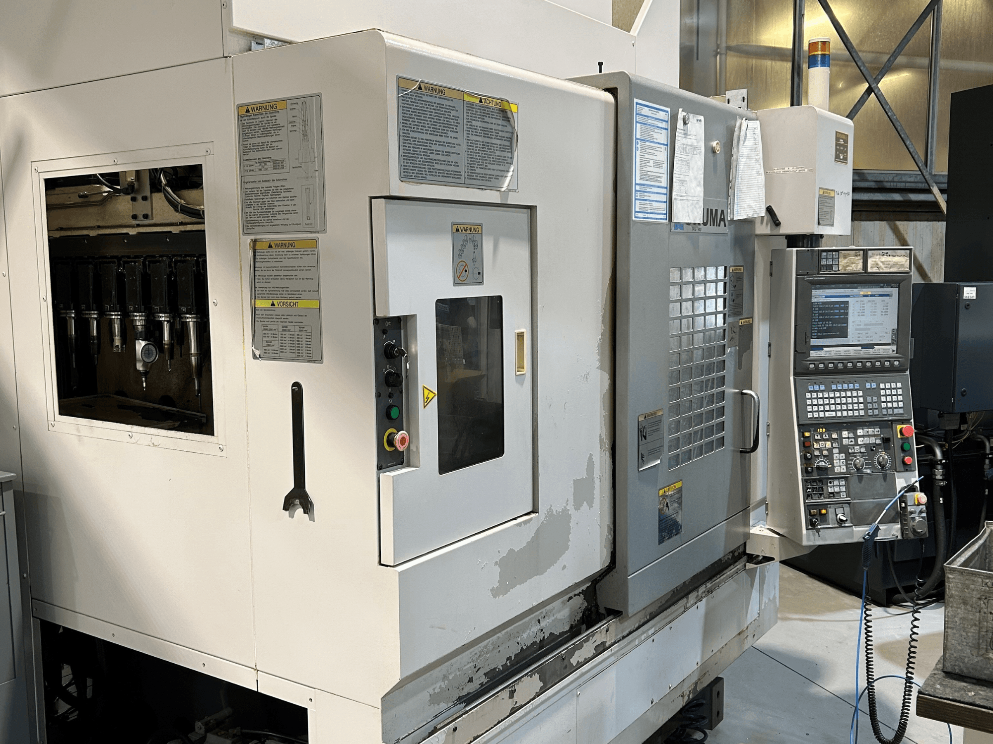 Widok z przodu maszyny CNC UMA z widocznymi uchwytami narzędzi, panelem sterowania, etykietami ostrzegawczymi i przyciskami operacyjnymi.