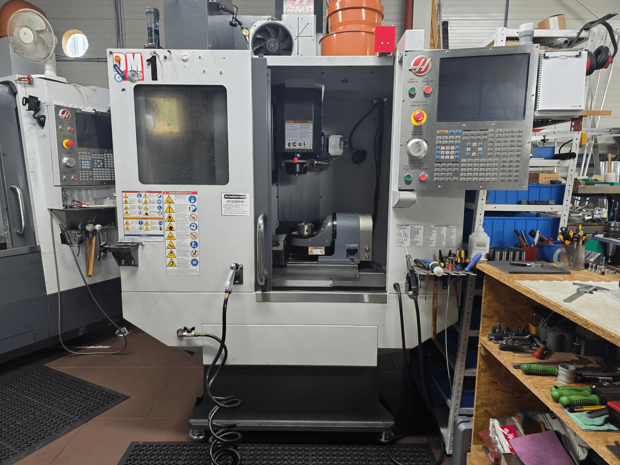 Maszyna CNC Haas model VF-2, widok z przodu, pokazujący elementy sterujące, obszar wrzeciona, etykiety bezpieczeństwa i narzędzia na pobliskim stole warsztatowym.