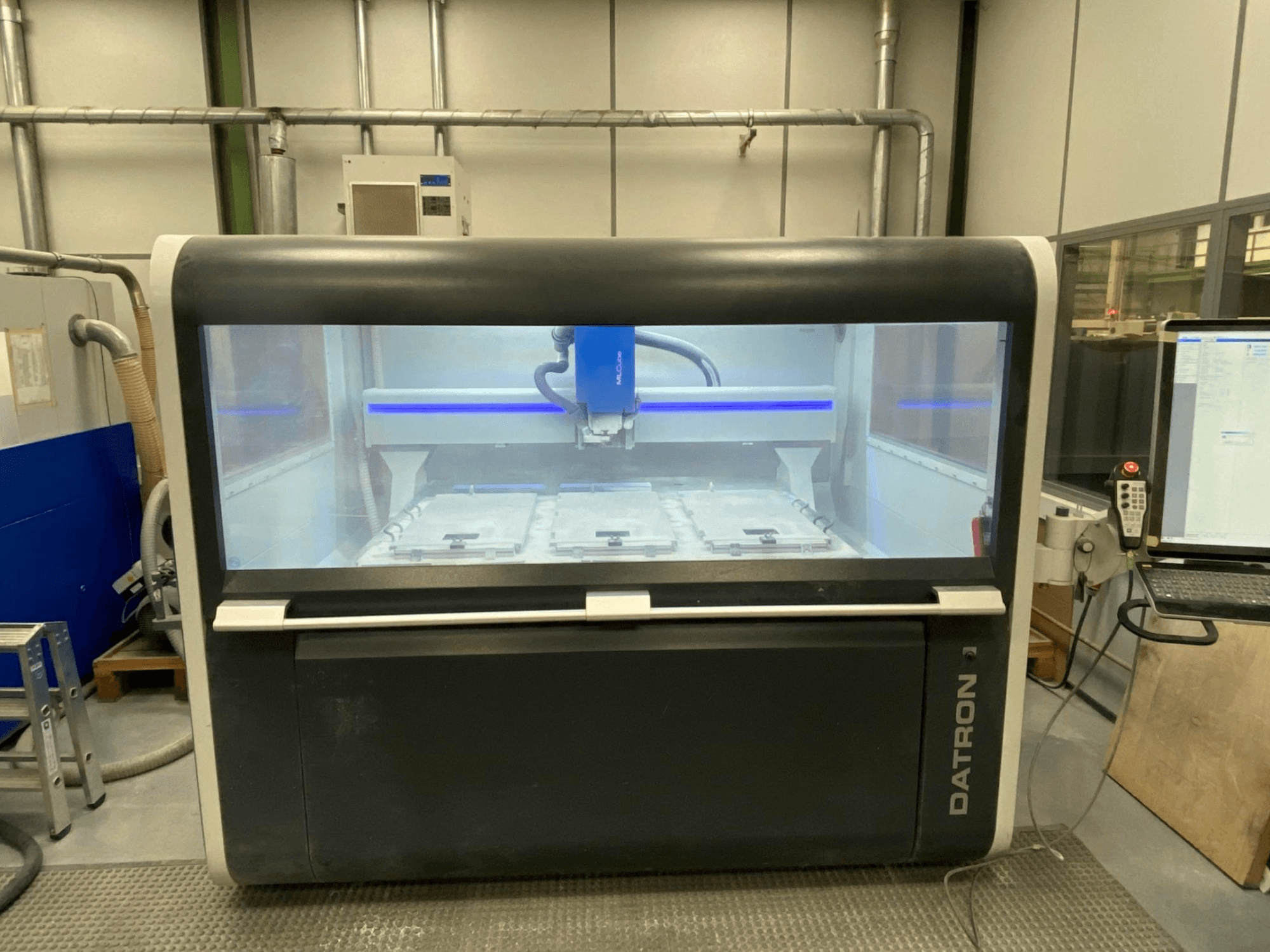 Widok z przodu pionowego centrum obróbczego Datron MLCube B, przedstawiający panel sterowania, obszar roboczy i głowicę tnącą wewnątrz szklanej obudowy.