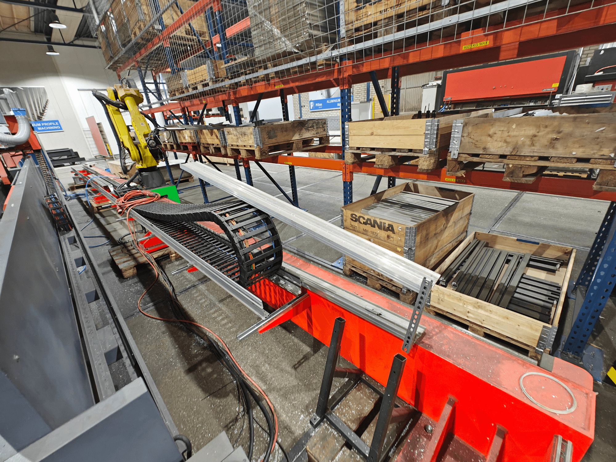 Ramię robota na stacji roboczej, widoczne pomarańczowe i czarne kable, ze skrzyniami Scania i profilami aluminiowymi w fabryce.