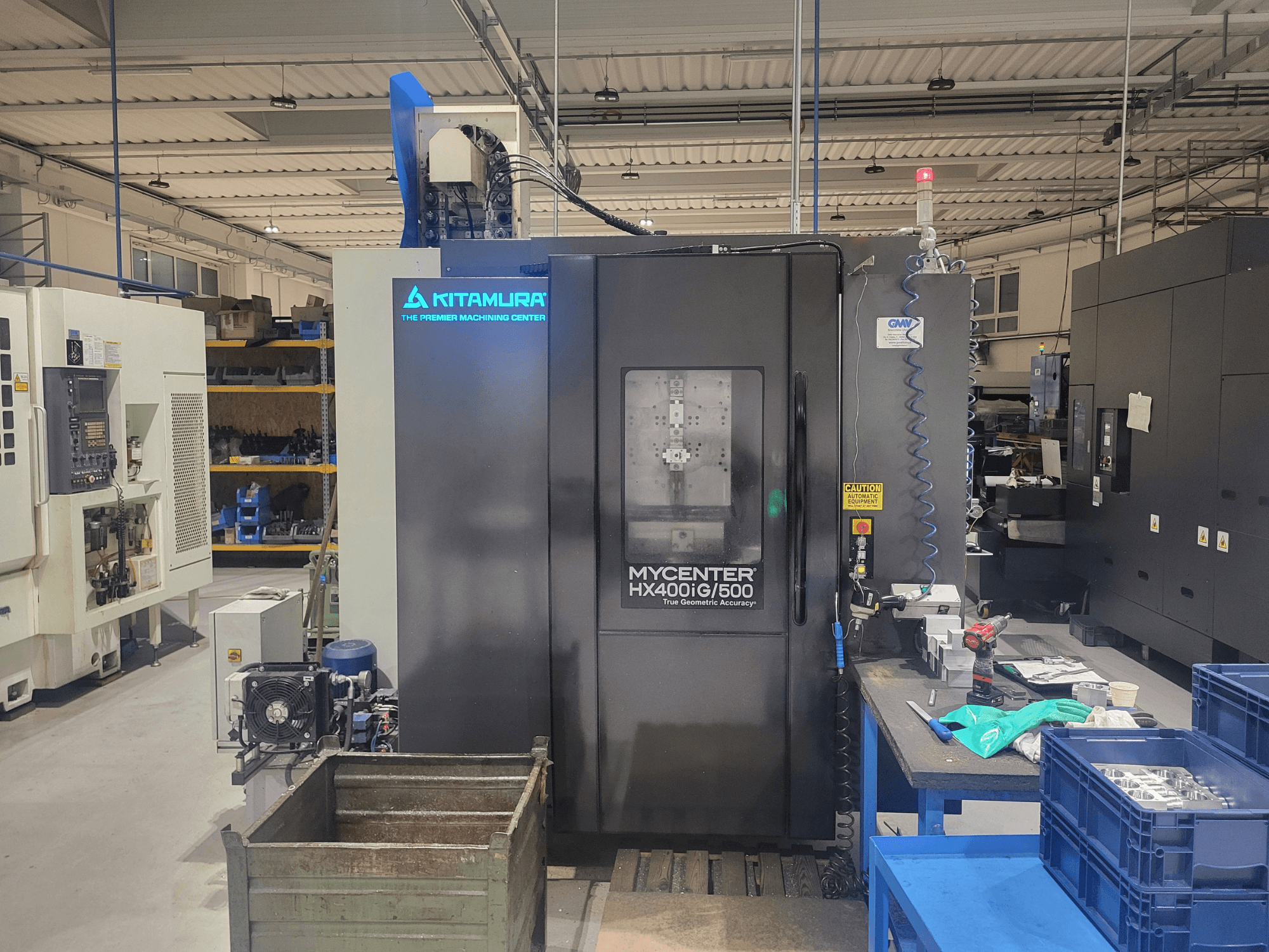 Maszyna CNC Kitamura Mycenter HX400iG/500 w warsztacie, widok z przodu, z elementami sterującymi, magazynem narzędzi i materiałami w pobliżu.
