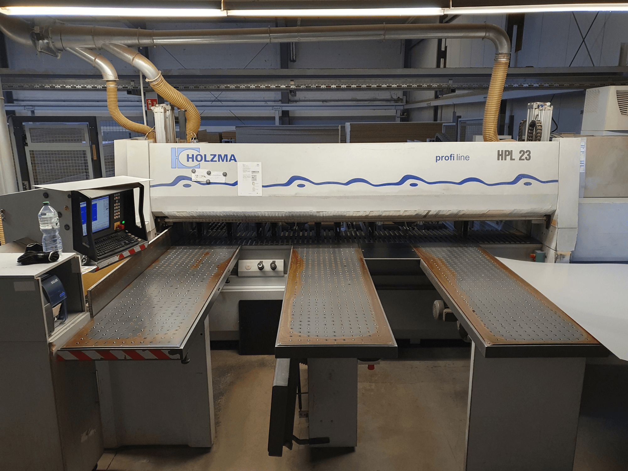 Maszyna do cięcia HOLZMA HPL 23 pokazana z przodu, z trzema perforowanymi stołami, terminalem komputerowym i butelką wody w pobliżu.