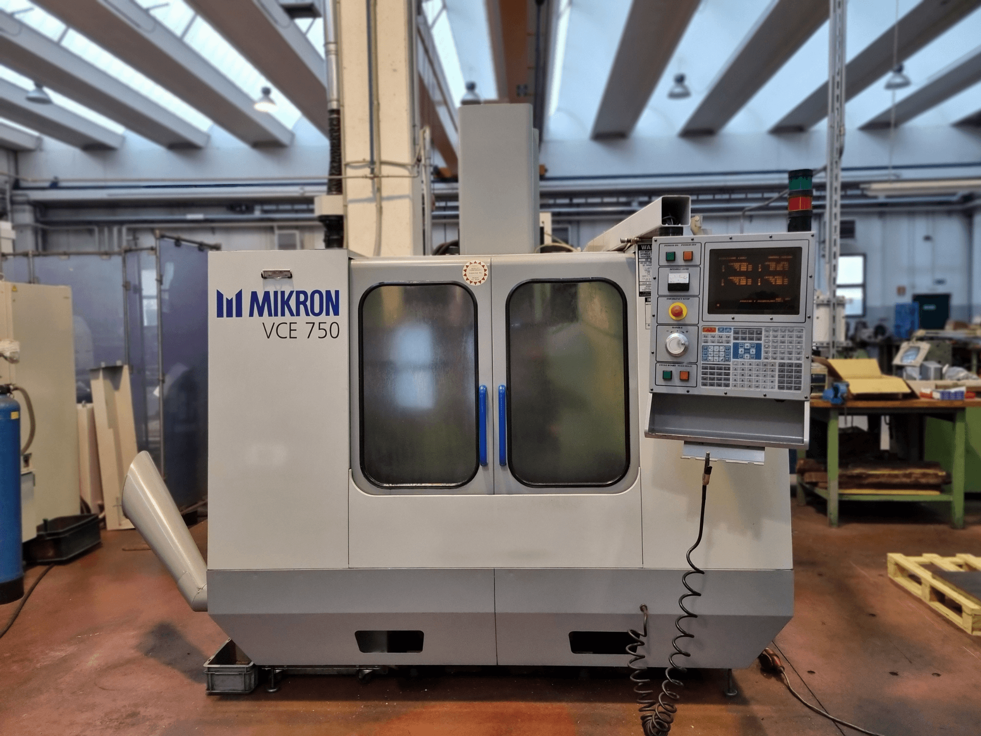 Biała maszyna CNC Mikron VCE 750 w warsztacie, widok z boku, z cyfrowym panelem sterowania i dużymi szklanymi drzwiami.