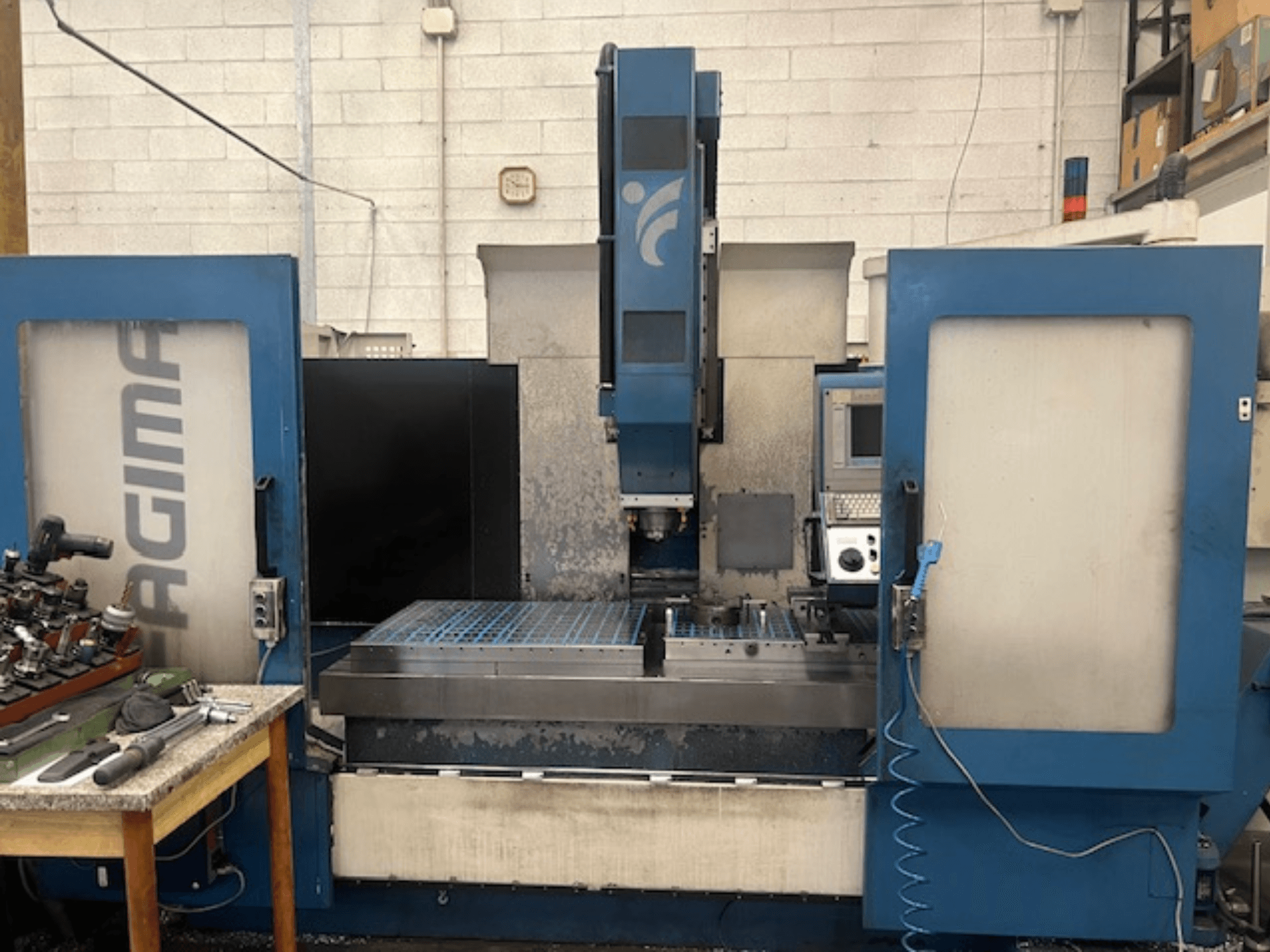Widok z przodu maszyny CNC JAGIMA z dużym metalowym obszarem roboczym, widocznym panelem sterowania i narzędziami na pobliskim stole.