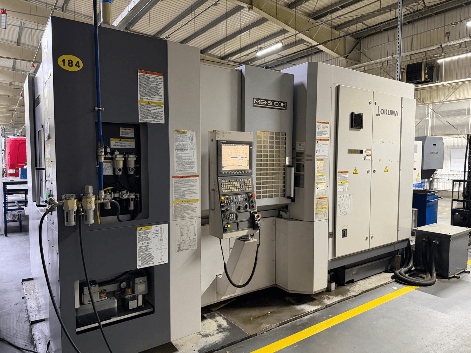 Widok z przodu poziomego centrum obróbczego Okuma MB 5000H z elementami sterującymi, etykietami i połączeniami hydraulicznymi.