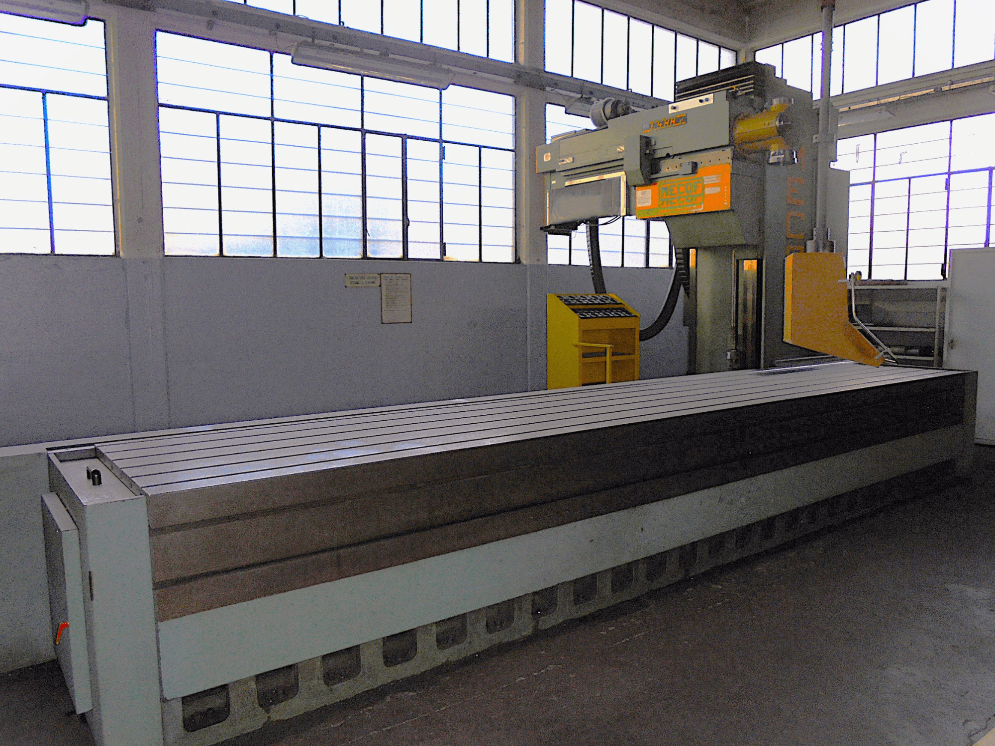 Nieznana marka, frezarka pionowa w fabryce, widok z boku, duży stół z rowkowaną powierzchnią i panelem sterowania.