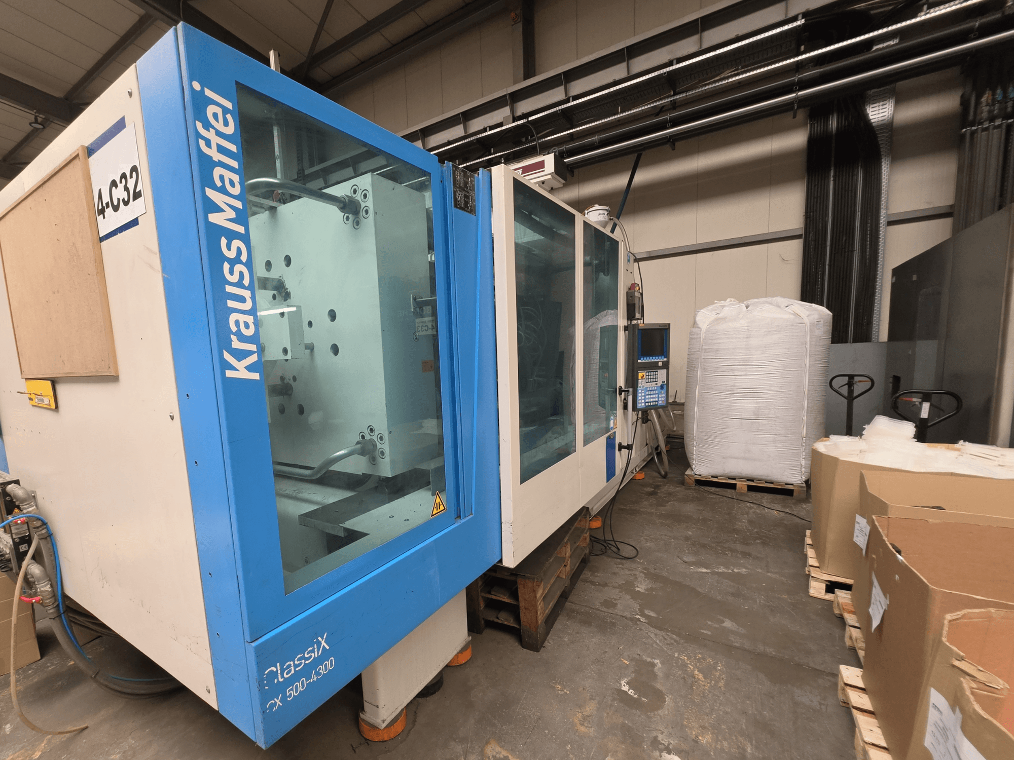 Hydrauliczna wtryskarka Krauss Maffei 500-4000 CX, widok z boku, panel sterowania i pobliskie materiały opakowaniowe.