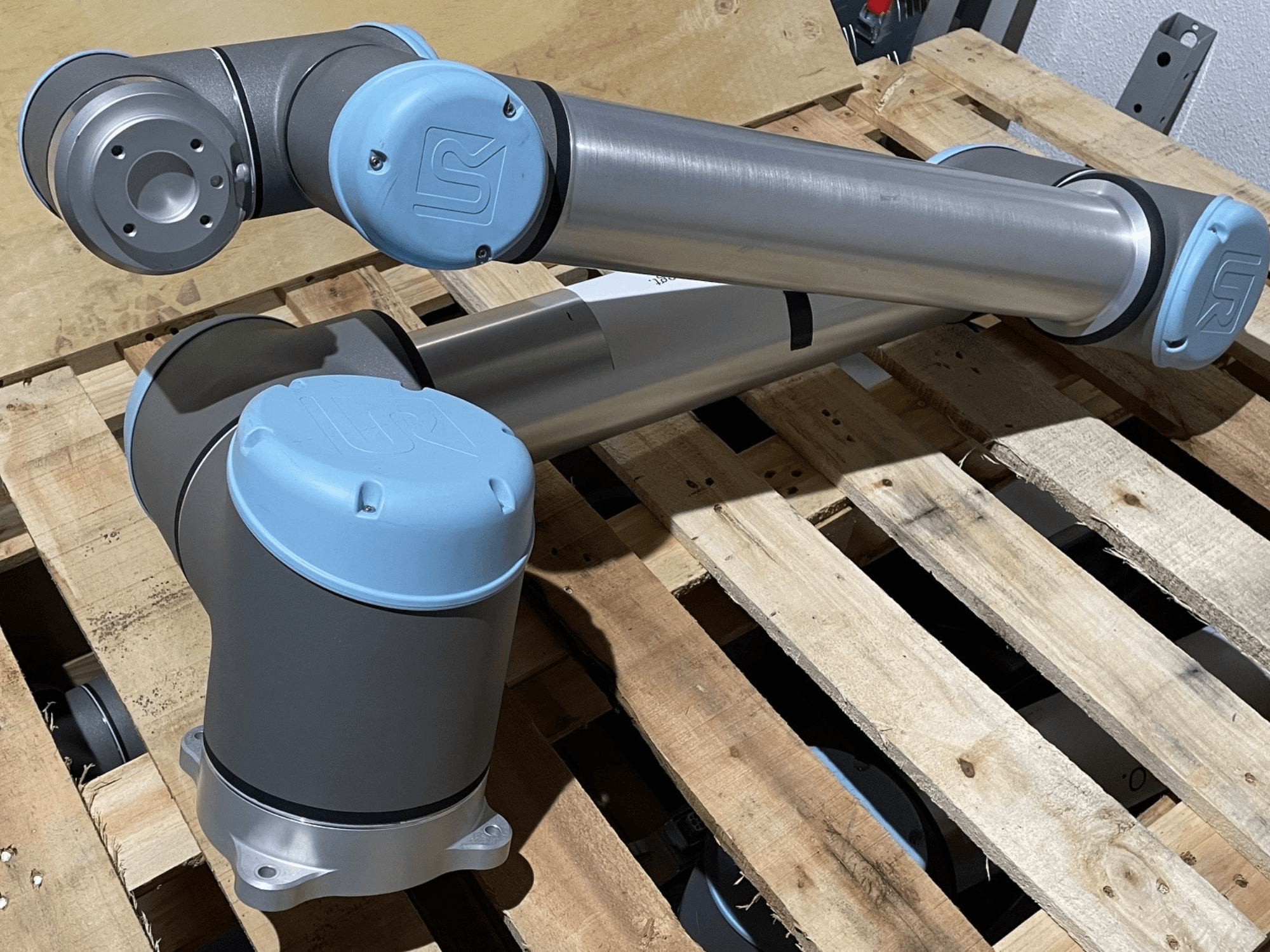 Srebrne ramię robota z niebieskimi zaślepkami, widok z przodu, prezentujące przeguby i mechaniczne detale, spoczywające na drewnianej palecie.