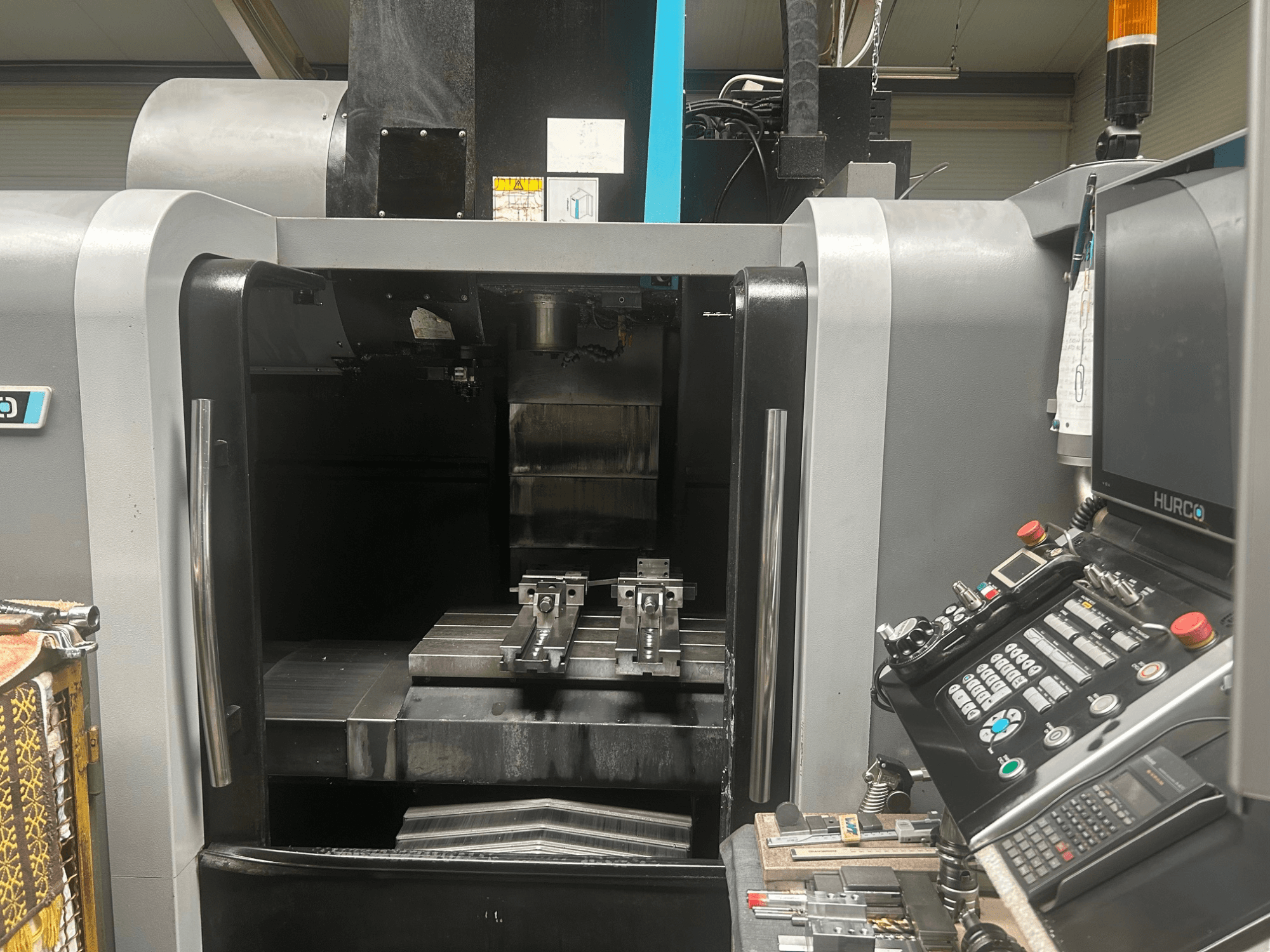 Widok z przodu centrum obróbczego CNC Hurco, pokazujący uchwyt narzędziowy, metalowy przedmiot obrabiany oraz panel sterowania z przyciskami i wyświetlaczem.