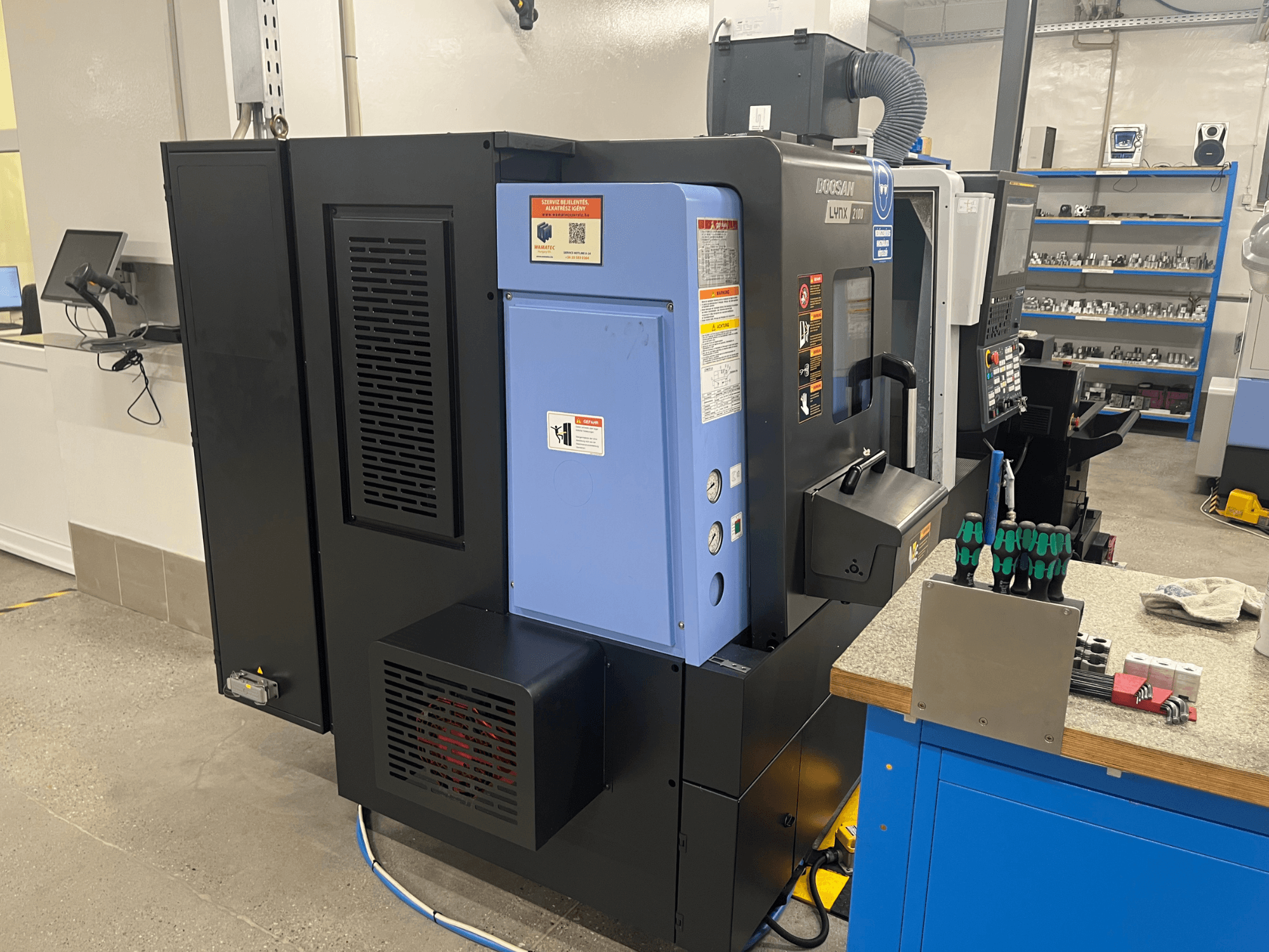 Tokarka Doosan Lynx 2100, widok z boku, panel sterowania, system wentylacji i narzędzia na stole warsztatowym.