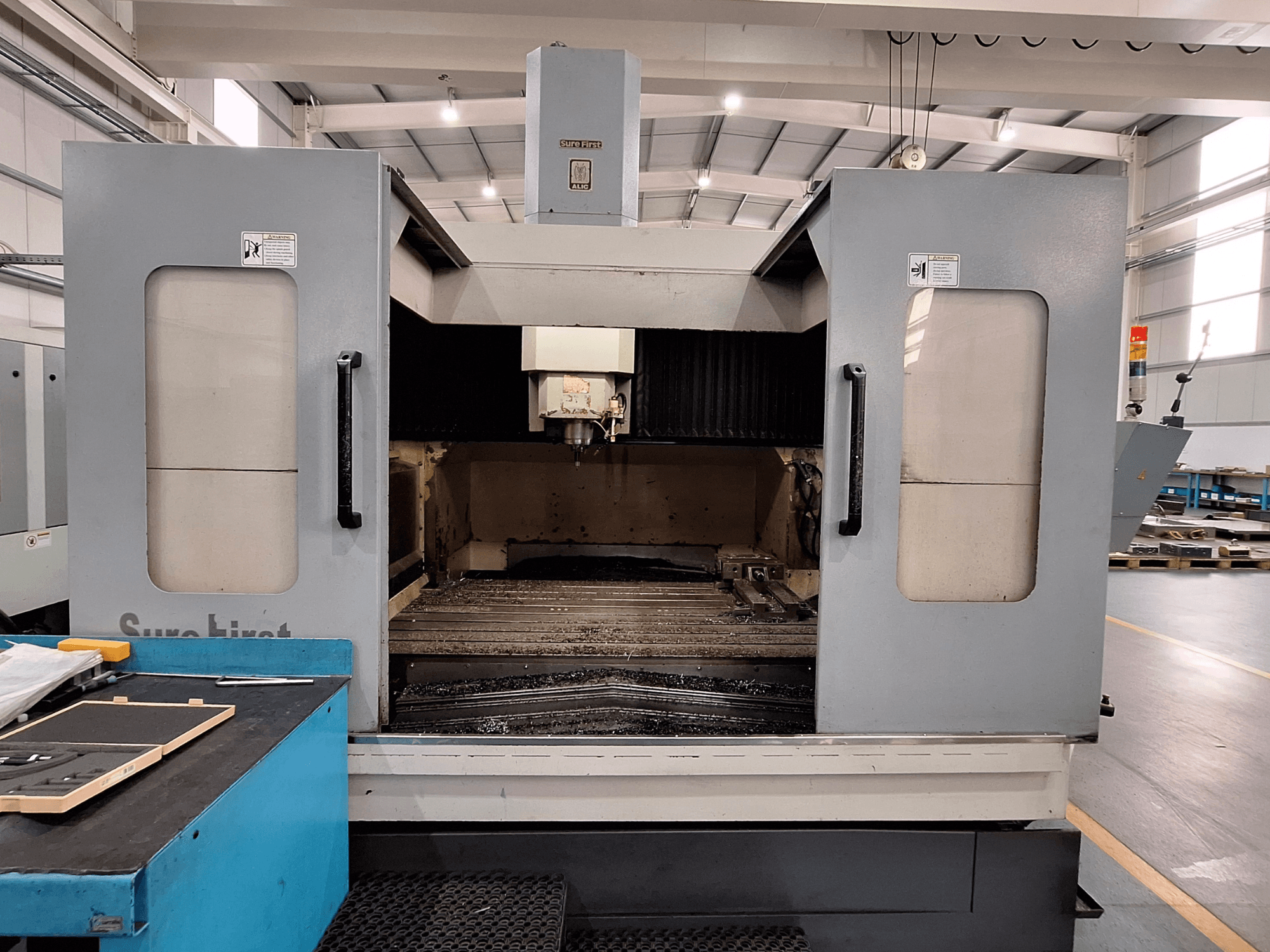 Pionowe centrum obróbcze Sure First QM-1836, widok z przodu pokazujący głowicę narzędziową i pusty obszar roboczy, nowoczesne otoczenie przemysłowe.