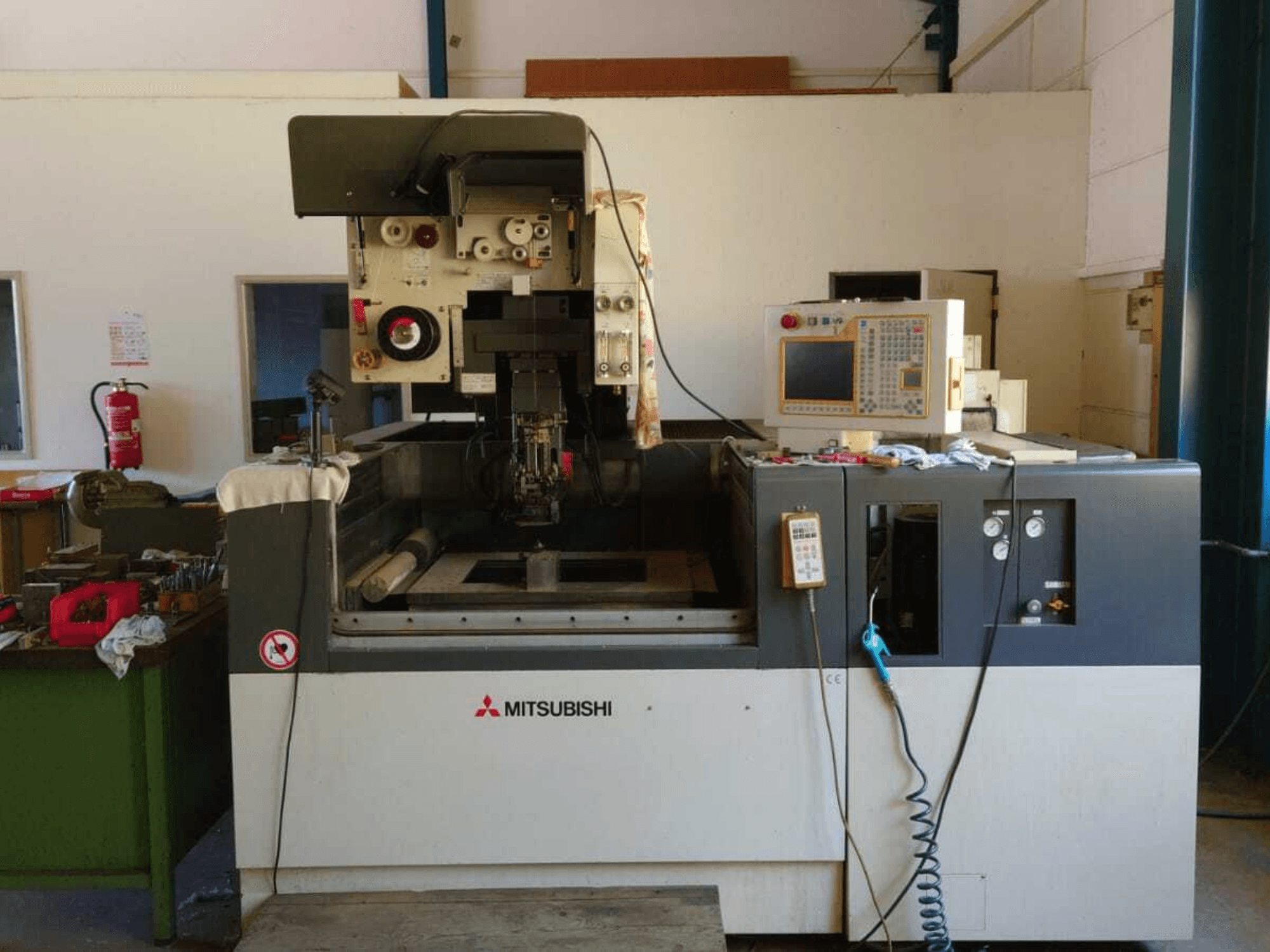 Widok z przodu maszyny do elektroerozji drutowej Mitsubishi FA20, na którym widoczny jest panel sterowania, obszar roboczy oraz oznakowanie bezpieczeństwa.