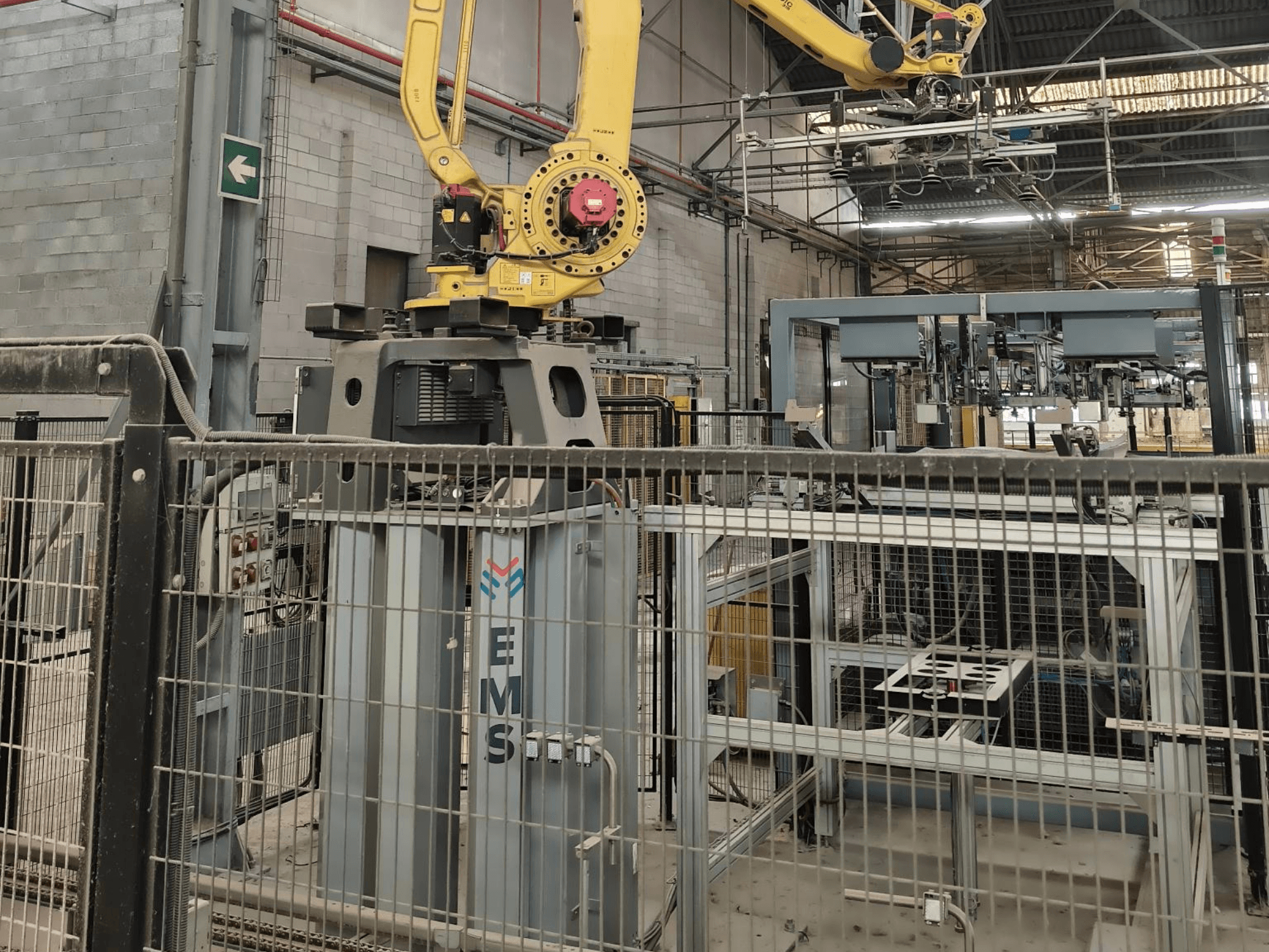 Widok z przodu ramienia robota FANUC M-410iC/315 umieszczonego nad narzędziem EMS, z wyposażeniem przemysłowym i ogrodzeniem w tle.