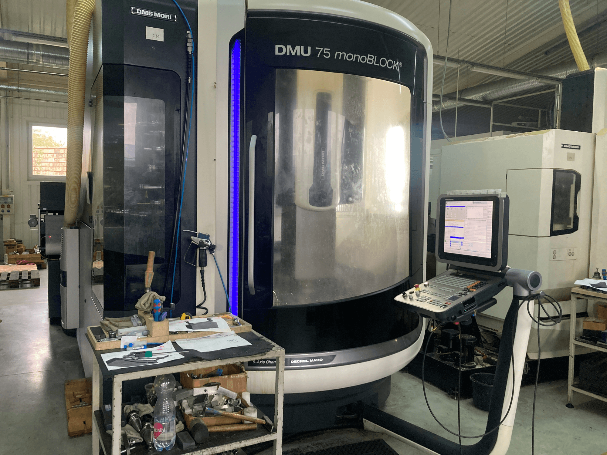 Maszyna CNC DMG MORI DMU 75 monoBLOCK®, widok z przodu, z panelem sterowania, narzędziami i obszarem roboczym z materiałami.