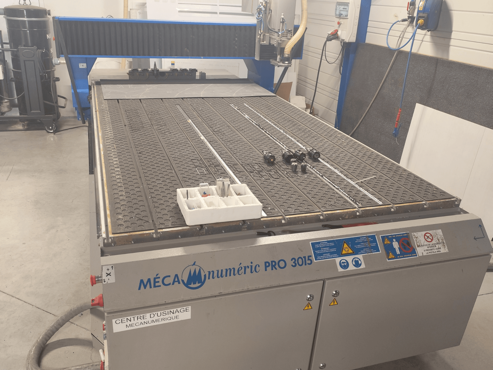 Widok z przodu frezarki CNC MECANUMERIC Pro 3015, przedstawiający szczegółowo stoł roboczy, panel sterowania oraz akcesoria umieszczone na stole.