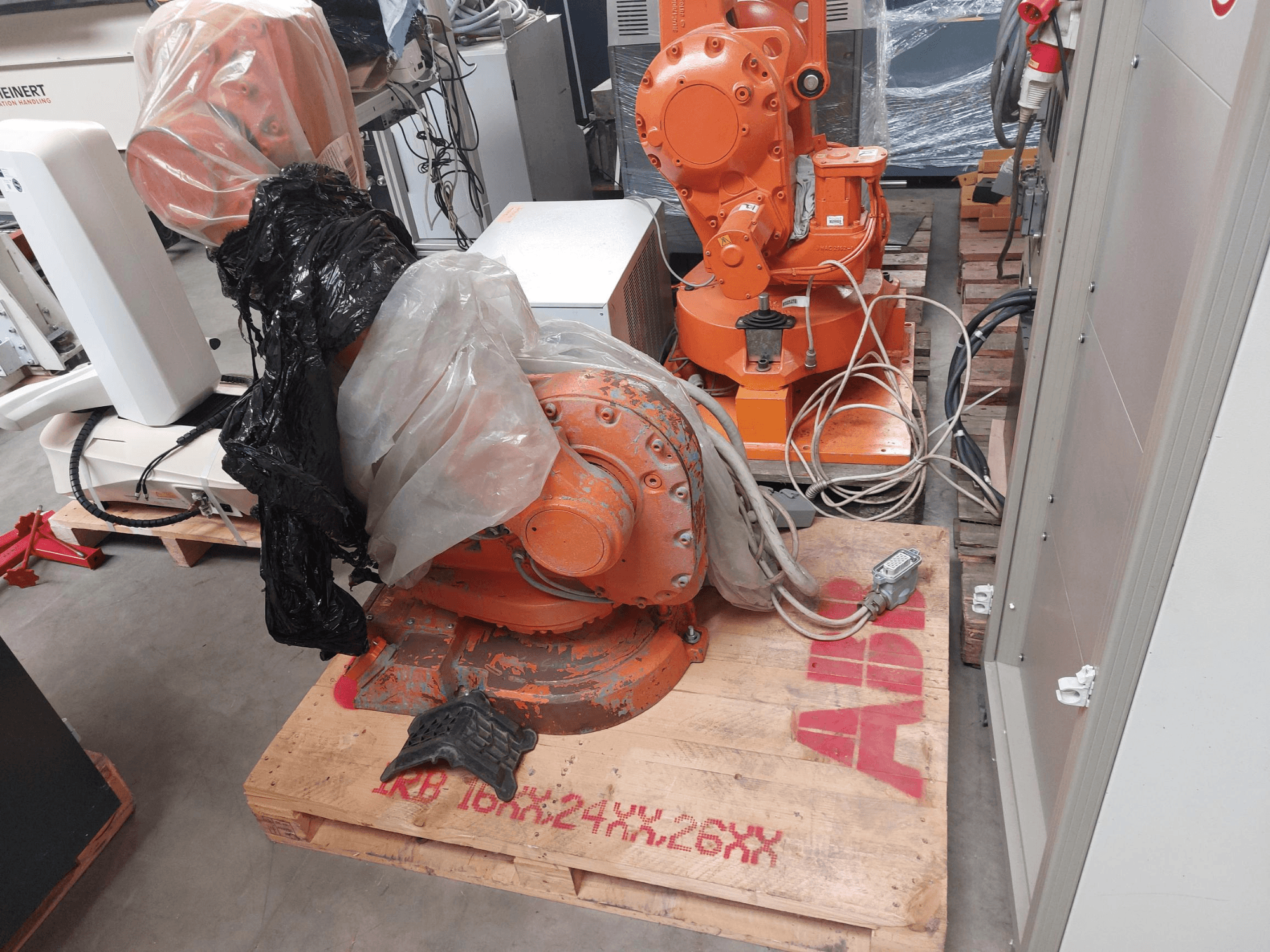 Ramię robota ABB IRB 1600 M2004 częściowo pokryte plastikiem, widziane z przodu, z kablami i dodatkowymi maszynami w pobliżu.