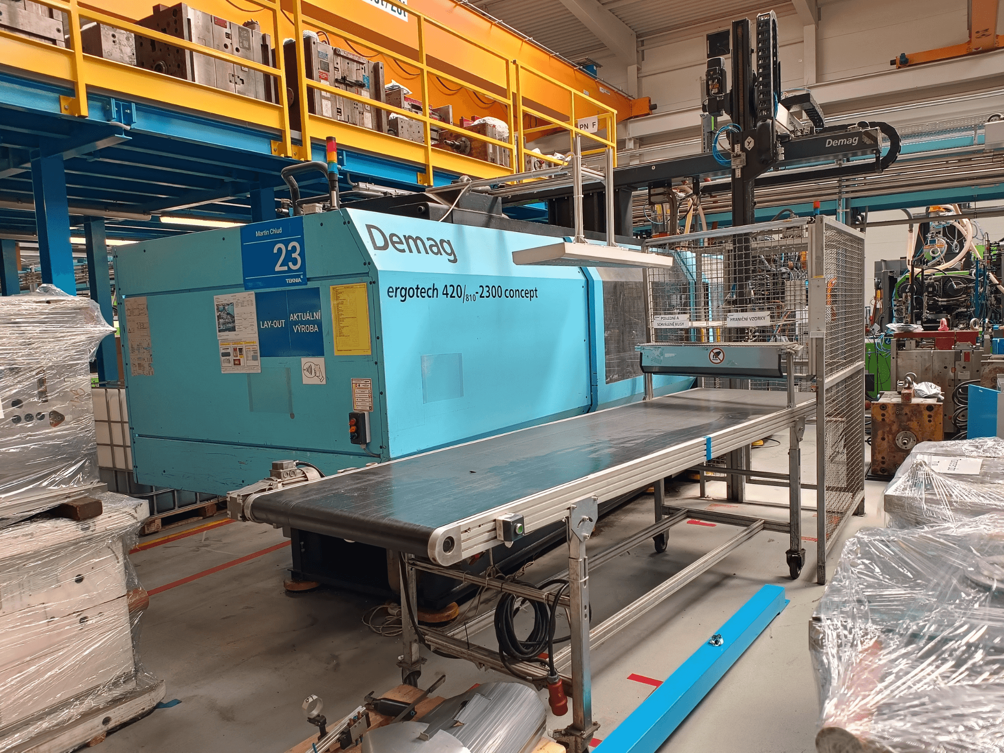 Hydrauliczna wtryskarka Demag ergotech 420/810 - 2300 w widoku z boku, z przenośnikiem i obudową bezpieczeństwa.