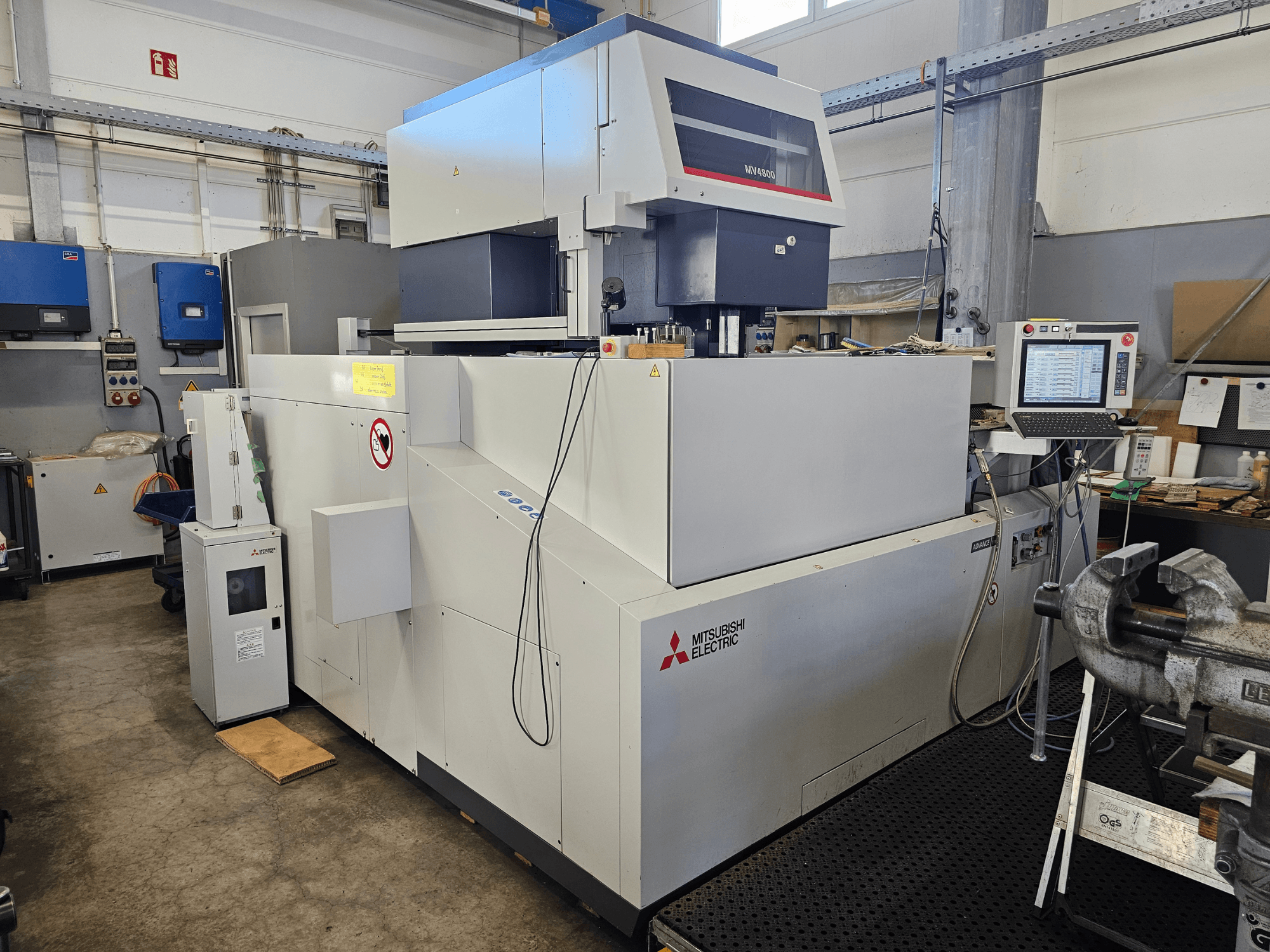 Elektrodrążarka drutowa Mitsubishi MV4800 w warsztacie, widok z boku pokazujący panel sterowania, etykiety bezpieczeństwa i otaczający sprzęt.