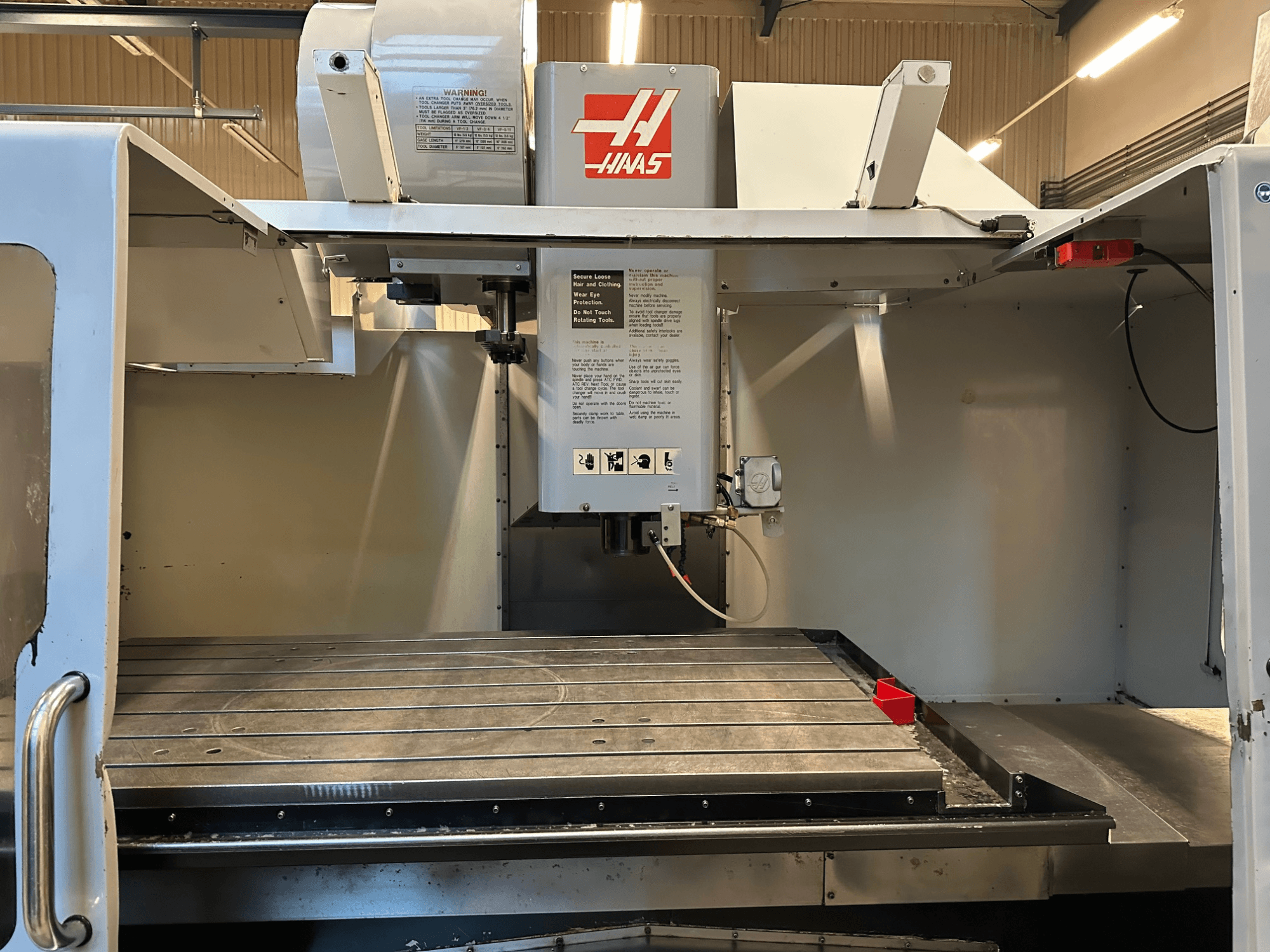 Frezarka CNC Haas, model VF-2, widok z przodu przedstawiający elementy sterujące, metalowy stół roboczy i etykietę bezpieczeństwa na panelu.