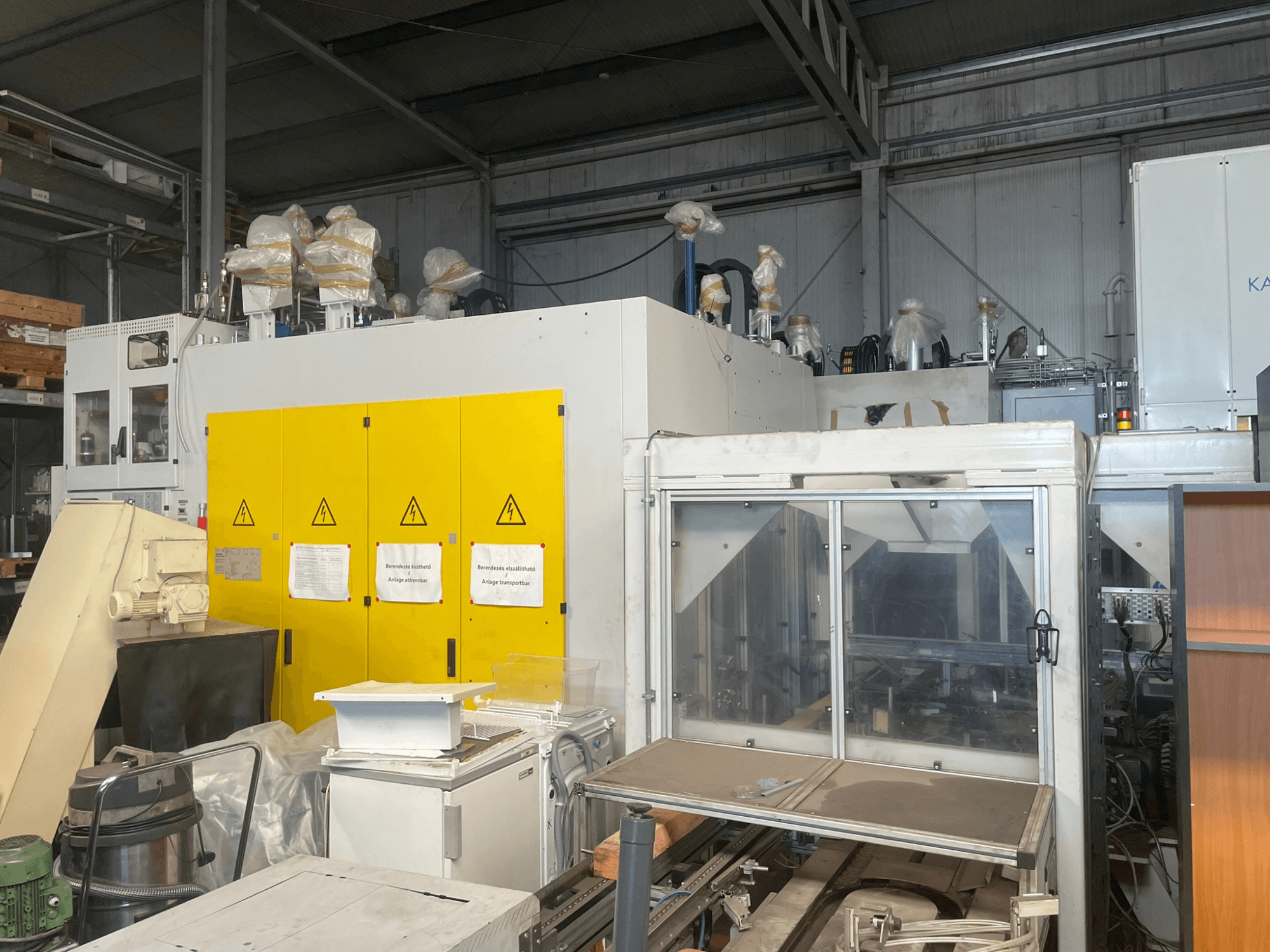 Maszyna przemysłowa z żółtym panelem sterowania, widok z boku, z etykietami ostrzegawczymi i plastikowymi osłonami, w otoczeniu magazynu.