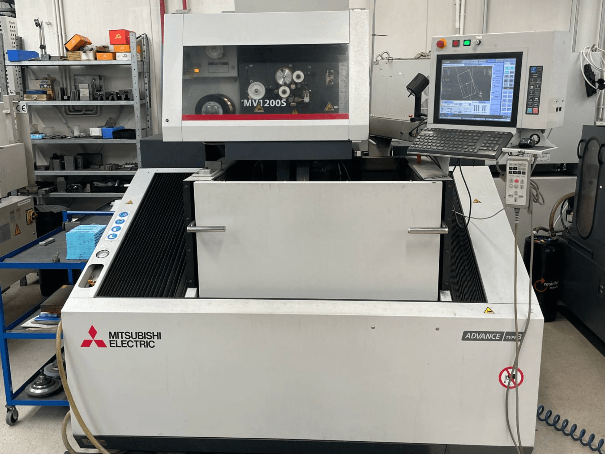 Maszyna do elektroerozji drutowej Mitsubishi MV1200S – widok z przodu, przedstawiający panel sterowania i obszar roboczy, z widocznymi ostrzeżeniami bezpieczeństwa.