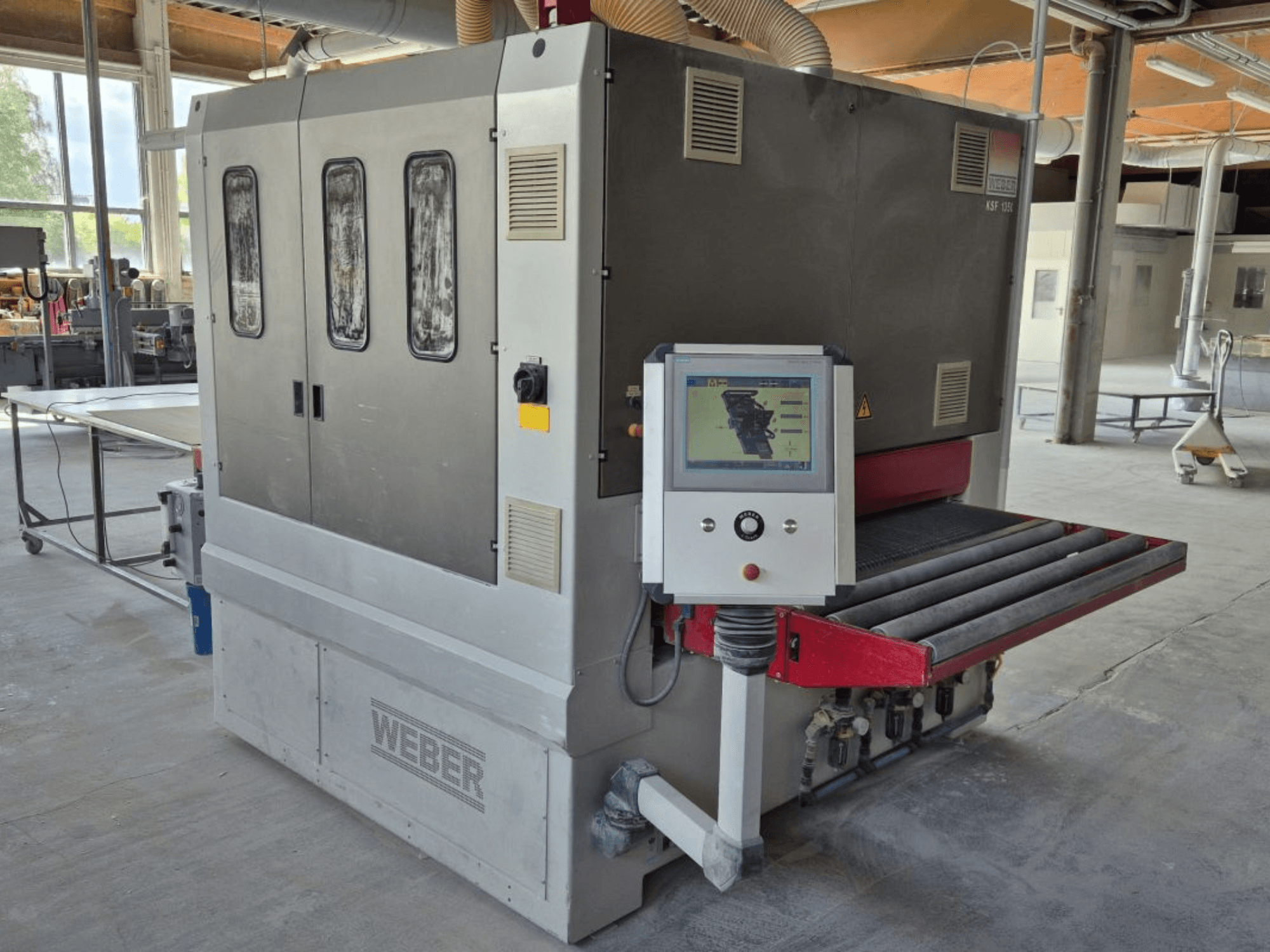Szlifierka taśmowa WEBER KSF-3-1350, widok z boku pokazujący elementy sterujące, panel zasilania i przenośnik szlifierski. Ustawienie przemysłowe z widocznymi maszynami.