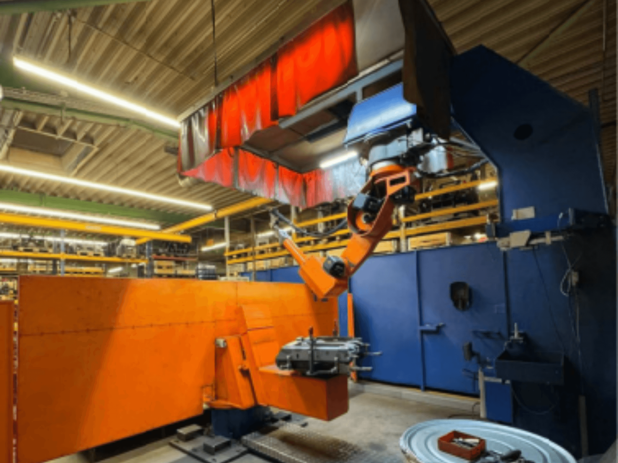 Robot spawalniczy CLOOS Romat 350 e w środowisku przemysłowym, widok z przodu z pomarańczowym ramieniem, trzymający obrabiany przedmiot pod kurtyną nad głową.