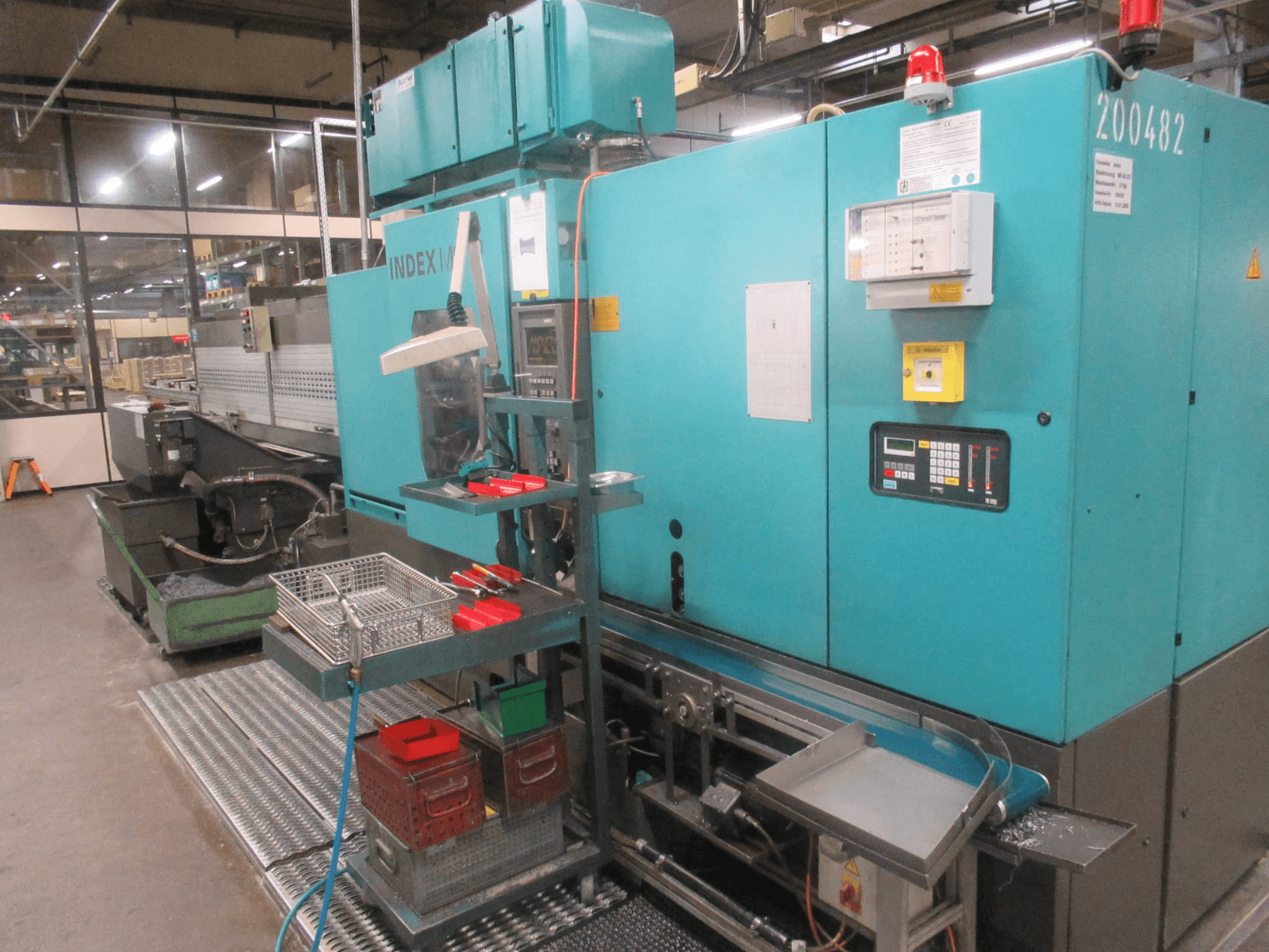 Turkusowa maszyna INDEX w fabryce, widok z boku; posiada panel sterowania, schowek na narzędzia i przenośnik. Widoczne pojemniki, węże i alarmy.