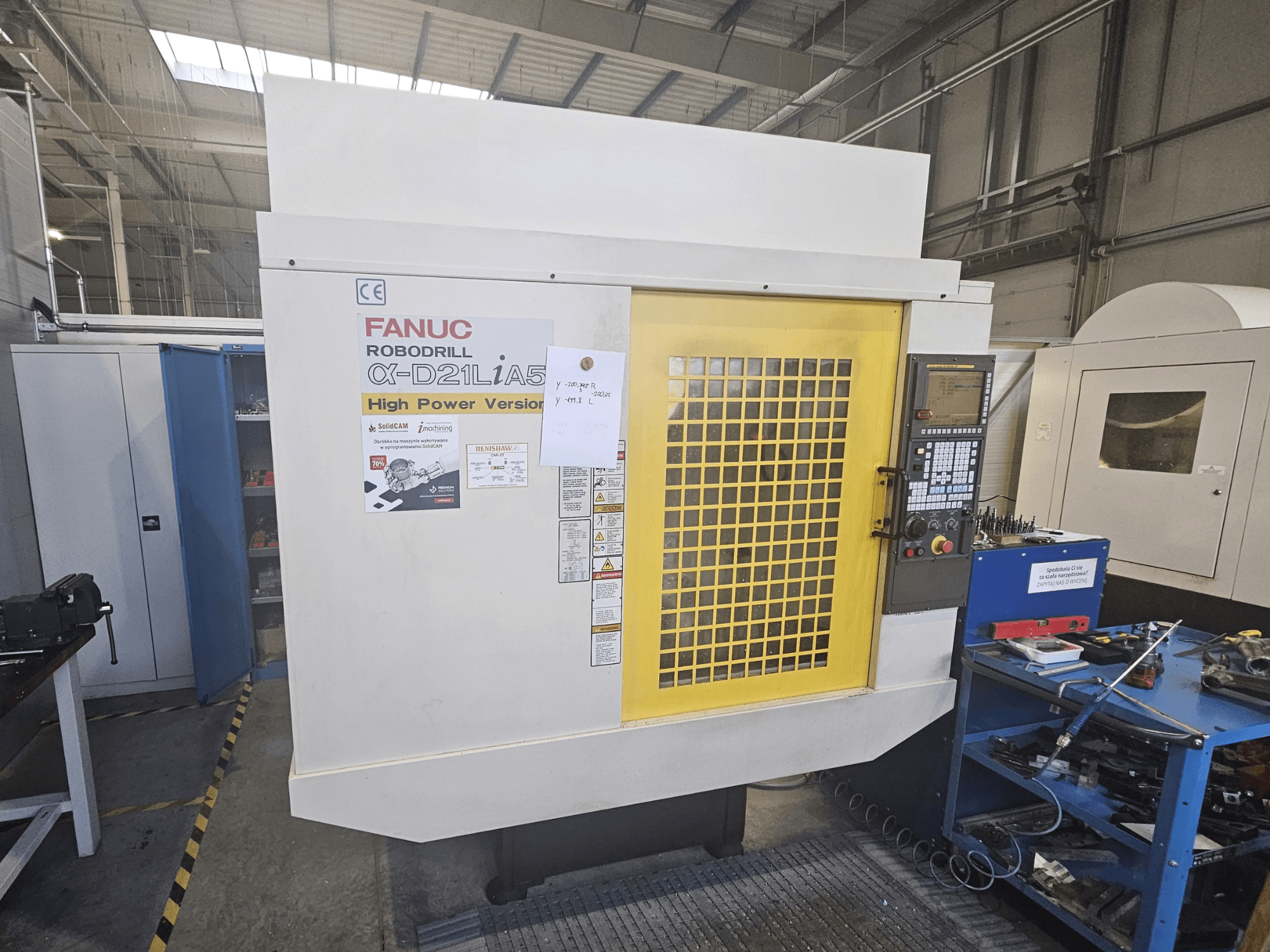 Widok z przodu wiertarki FANUC Robodrill α-D21LiA5 z żółtymi drzwiczkami bezpieczeństwa, panelem sterowania i narzędziami w pobliżu.