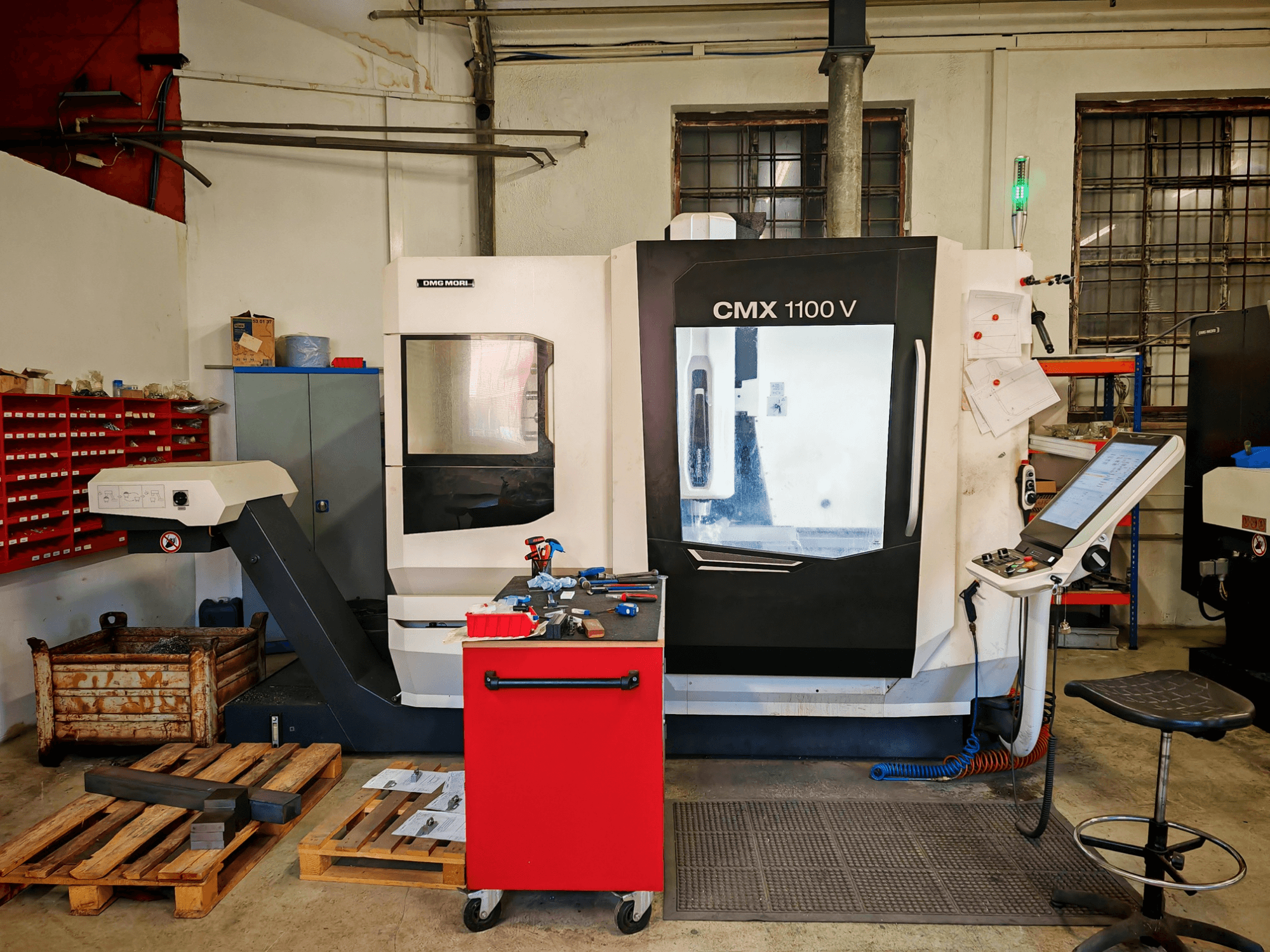Pionowe centrum obróbcze DMG MORI CMX 1100 V w warsztacie, widok z przodu, przedstawiający panel sterowania i otaczające go narzędzia.