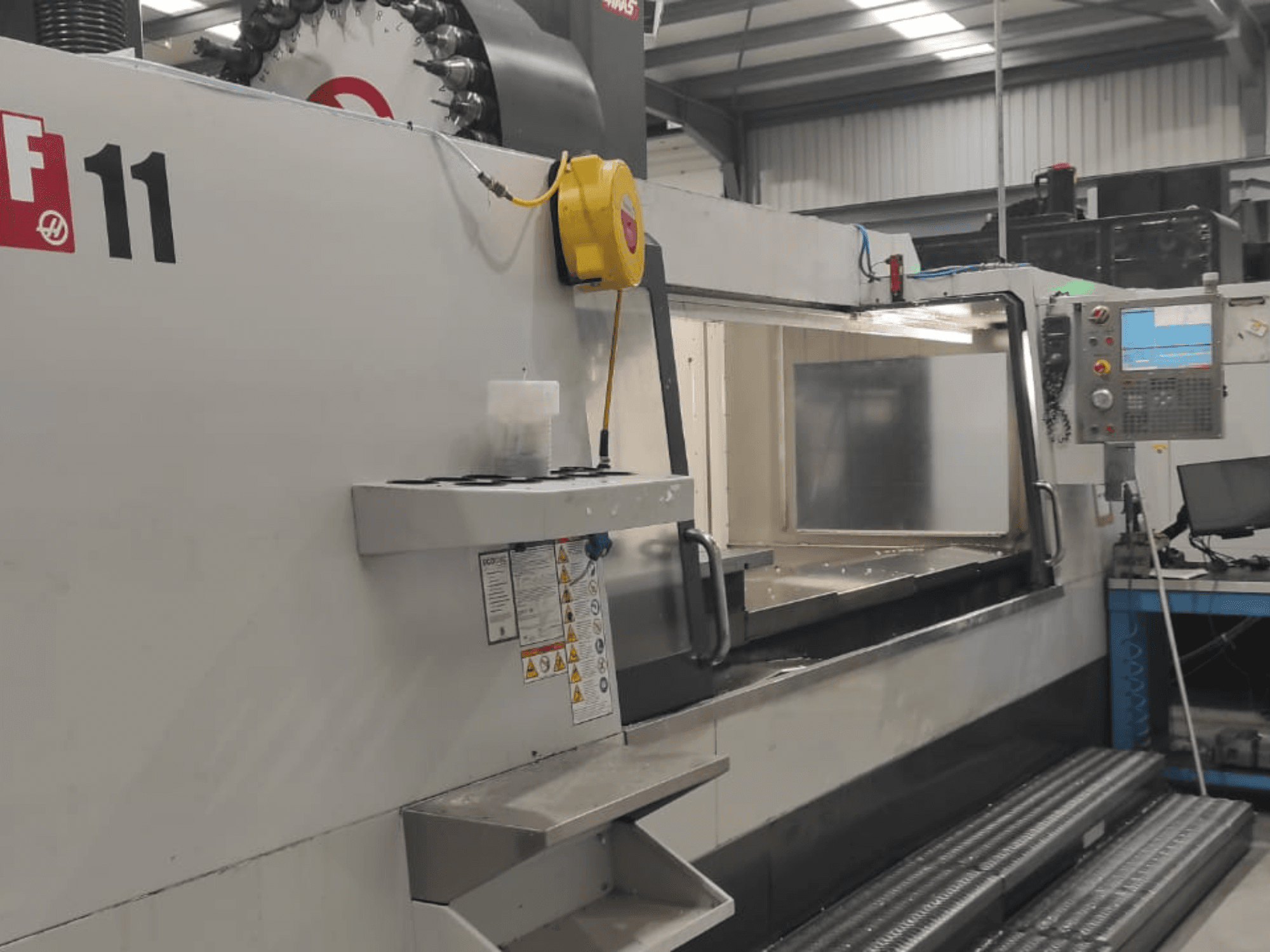 Maszyna firmy Haas, model VF-11, pokazana z przodu z widocznym panelem sterowania, dużym obszarem roboczym i etykietami bezpieczeństwa.