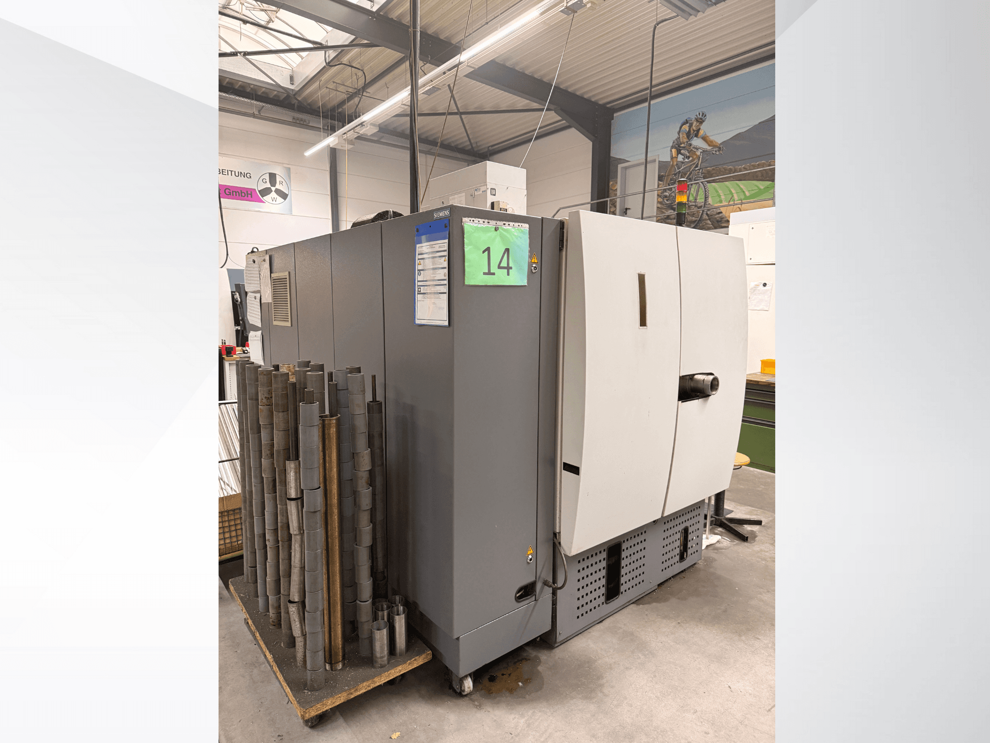 Szara maszyna przemysłowa firmy Siemens, widok z boku, z magazynem cylindrycznych elementów metalowych obok i widocznymi etykietami wyposażenia.