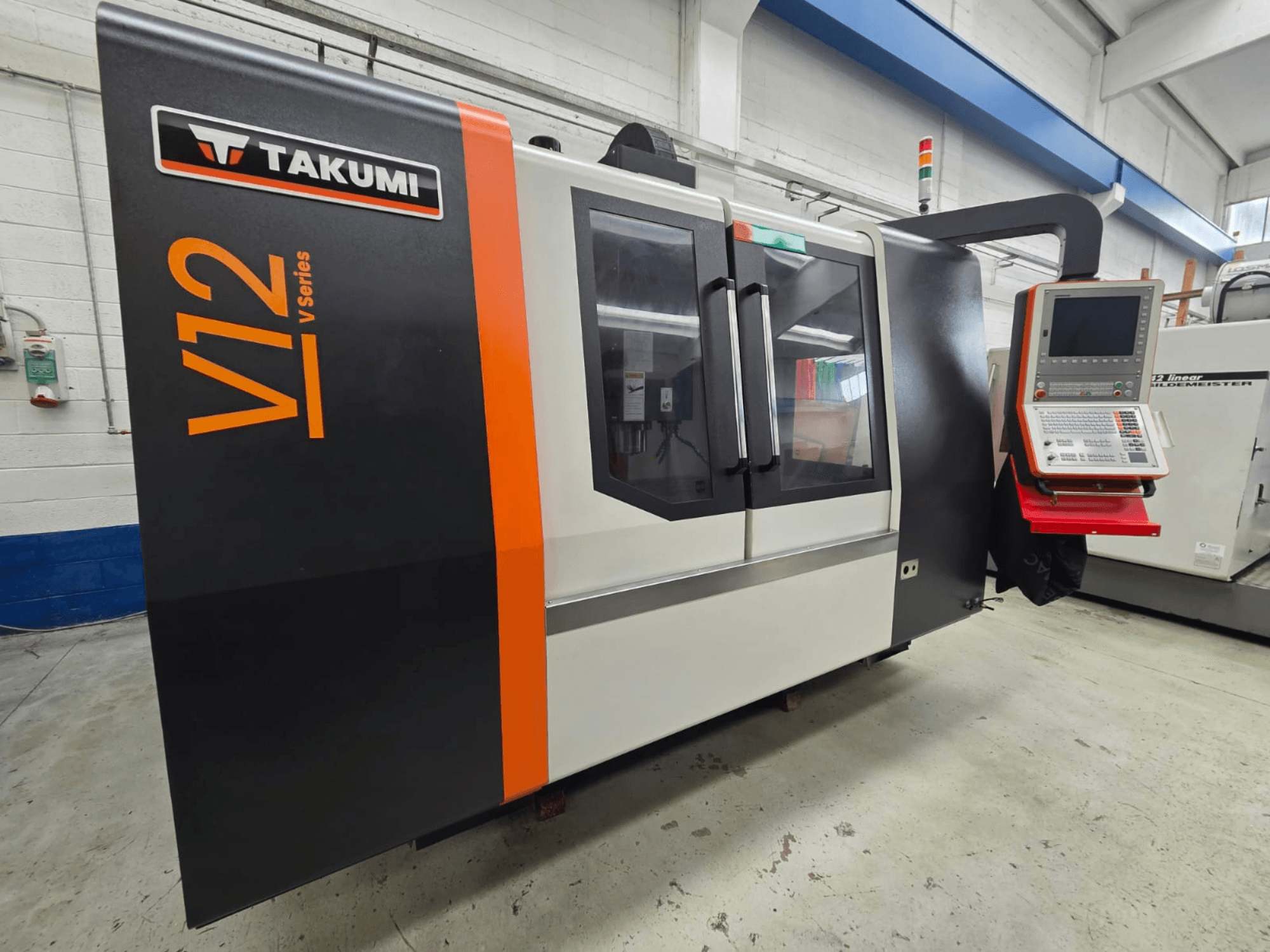 Frezarka CNC serii Takumi V12 V, widok z przodu, posiada panel sterowania i duże szklane okna.