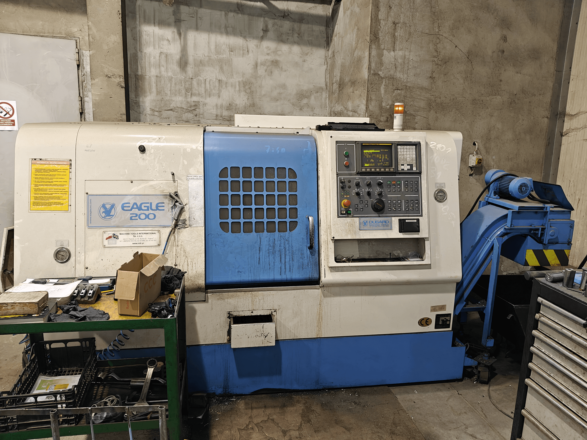 Maszyna CNC Eagle 200, widok z przodu z panelem sterowania, różne narzędzia na zielonym wózku. Widoczne zużycie i działające światła.