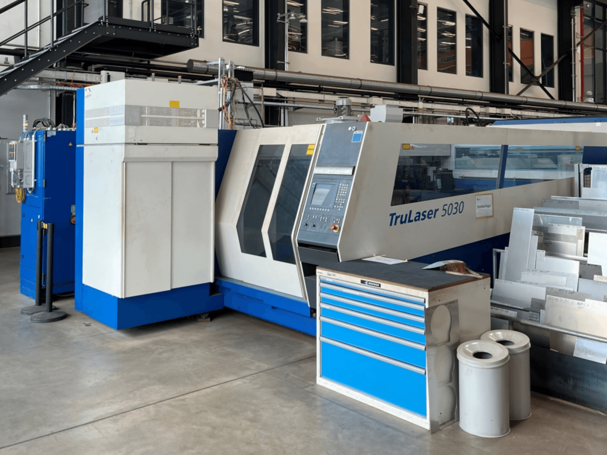 Maszyna do cięcia laserowego CO₂ TRUMPF TruLaser 5030 widziana z przodu, z panelem sterowania i znajdującym się obok stołem roboczym z blachami.