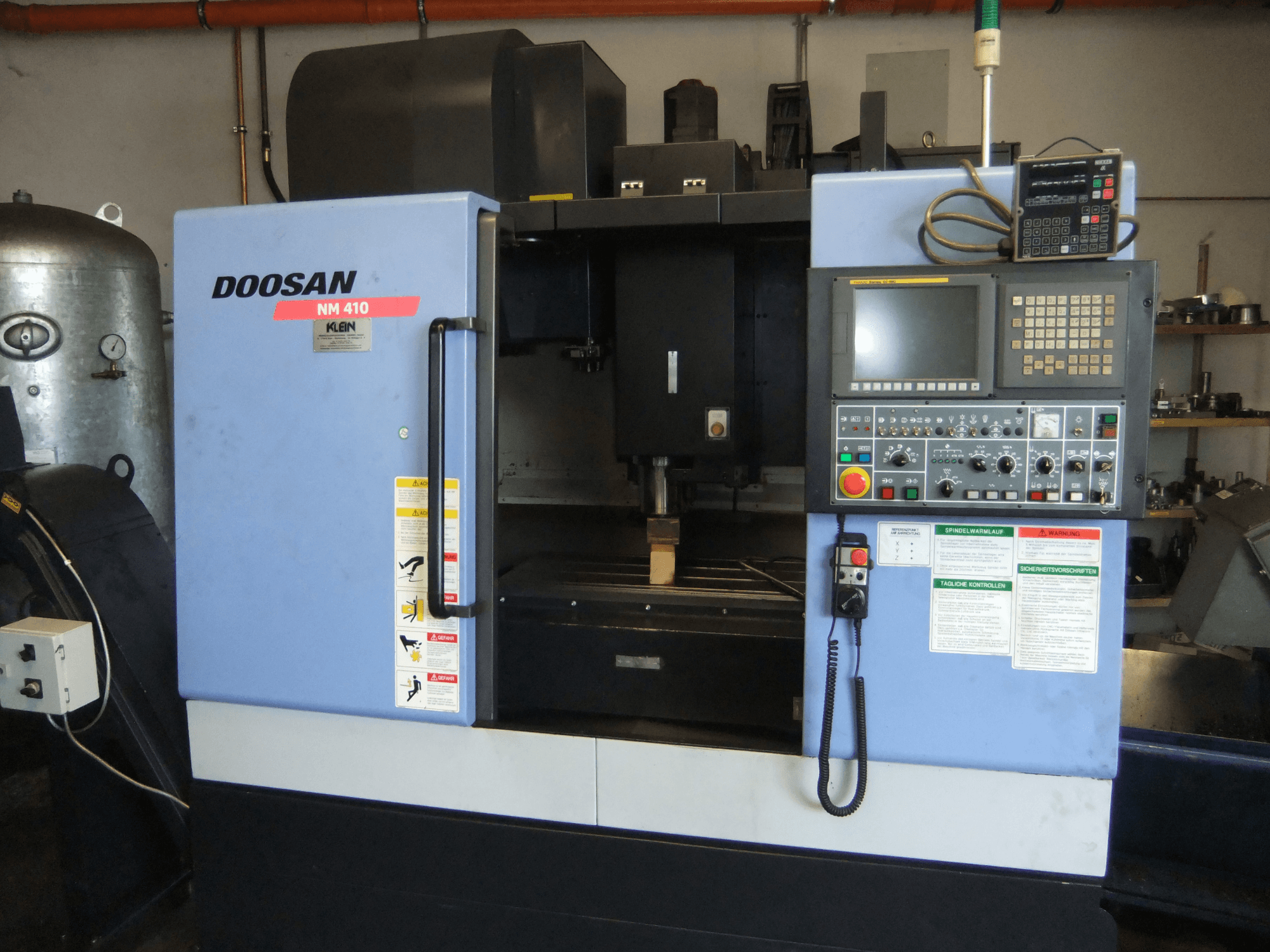 Pionowe centrum obróbcze DOOSAN NM 410 – widok z przodu, przedstawiający panel sterowania, obszar wrzeciona oraz etykiety bezpieczeństwa.