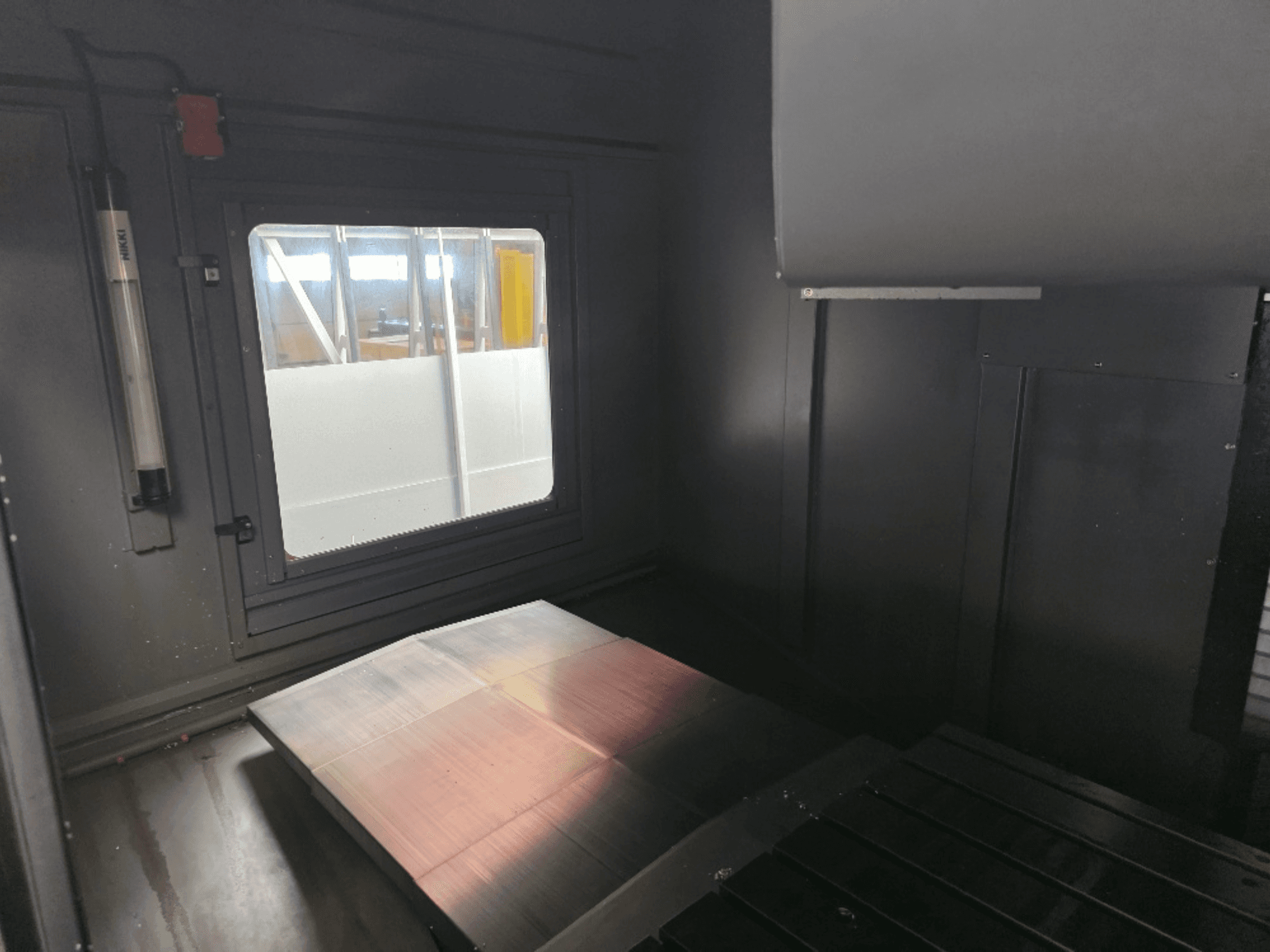 Widok wnętrza obudowy maszyny z polerowaną metalową płytą, bocznym oknem i minimalnym oświetleniem. Brak widocznej marki.