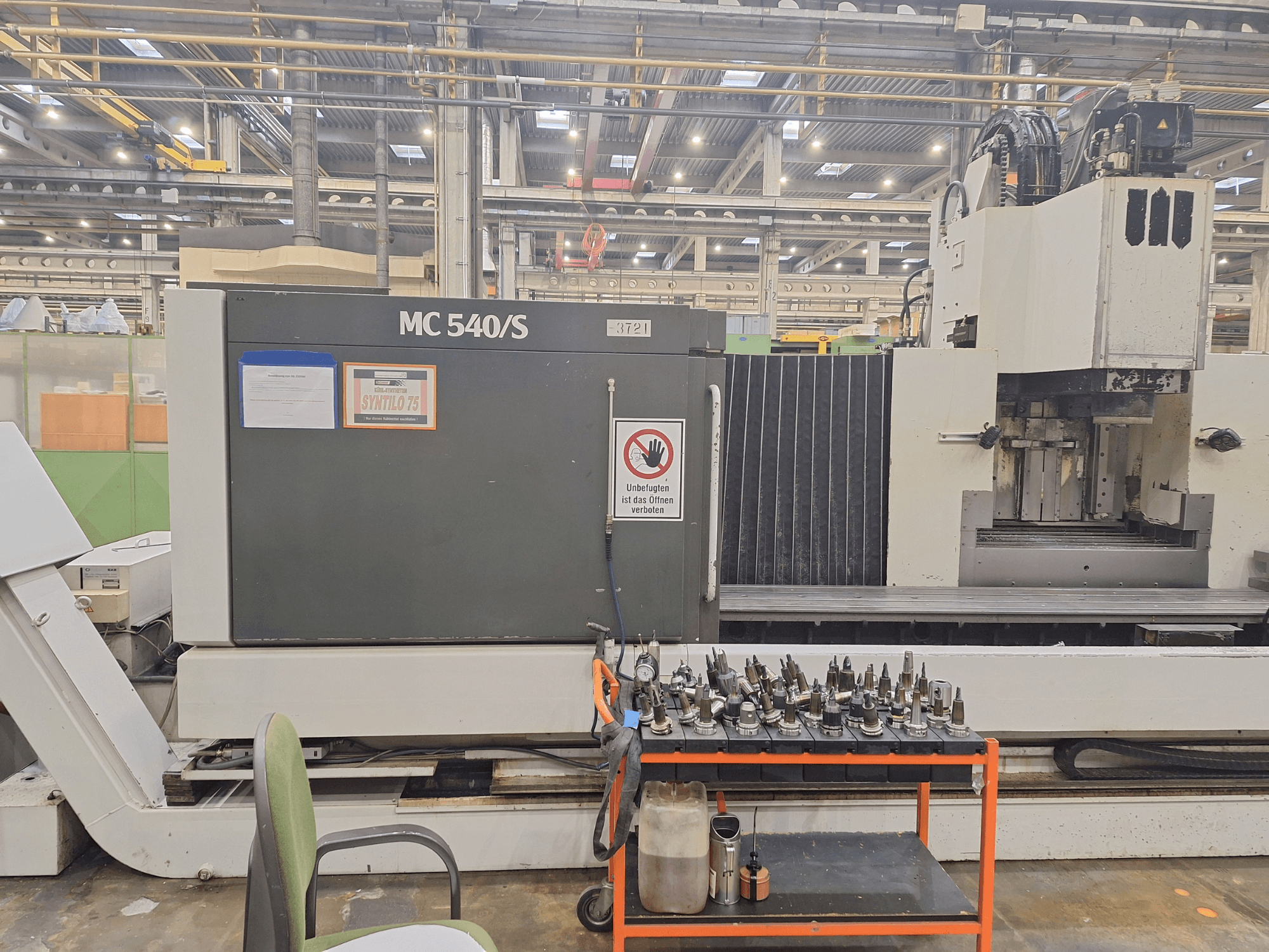 Pionowe centrum obróbcze STAMA MC 540S w warsztacie, widok z boku z uchwytami narzędziowymi na wózku obok.