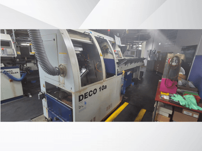 Maszyna CNC DECO 10a w warsztacie, widok z przodu; wyposażona w panel sterowania i system odsysania pyłu, w pobliżu stół warsztatowy.