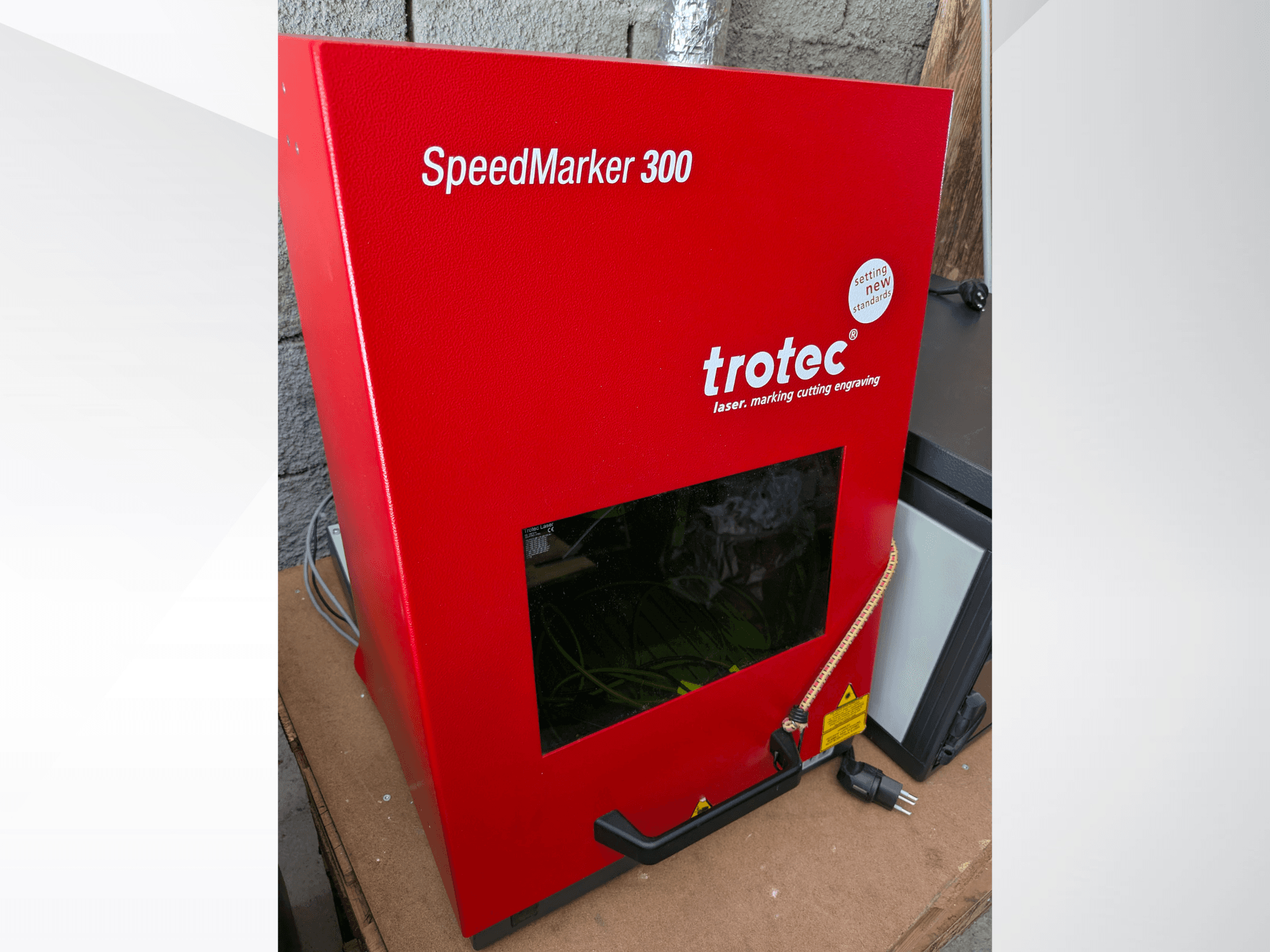 Czerwona znakowarka laserowa Trotec SpeedMarker 300 w widoku z przodu, wyposażona w duże okno podglądu i złącze zasilania.