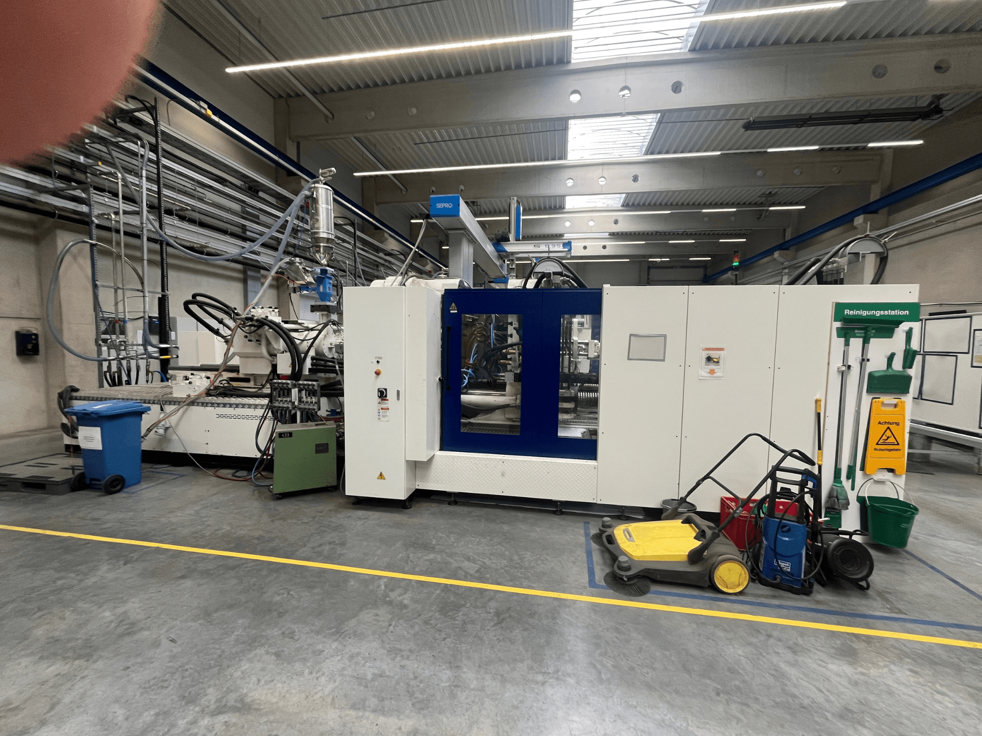 Maszyna przemysłowa w fabryce, widok z przodu, z niebieskimi i białymi panelami, wężami i sprzętem czyszczącym w pobliżu.