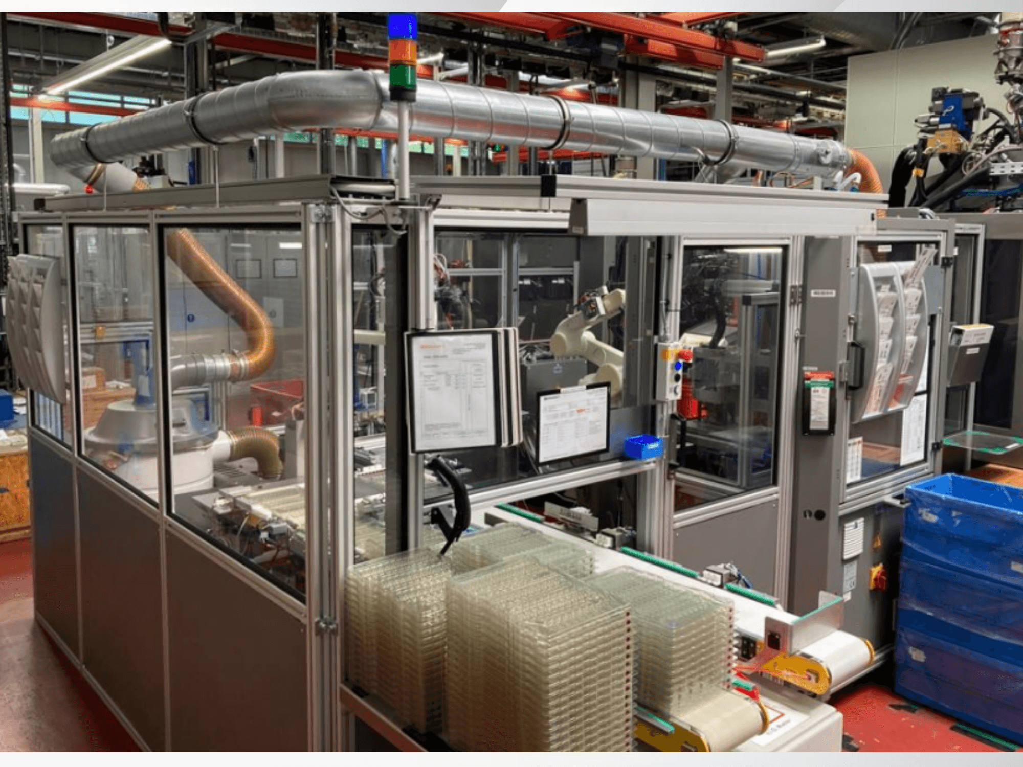 Widok z przodu zautomatyzowanej maszyny z metalowymi rurami, panelem sterowania i ułożonymi w stos przezroczystymi plastikowymi pojemnikami na hali produkcyjnej.