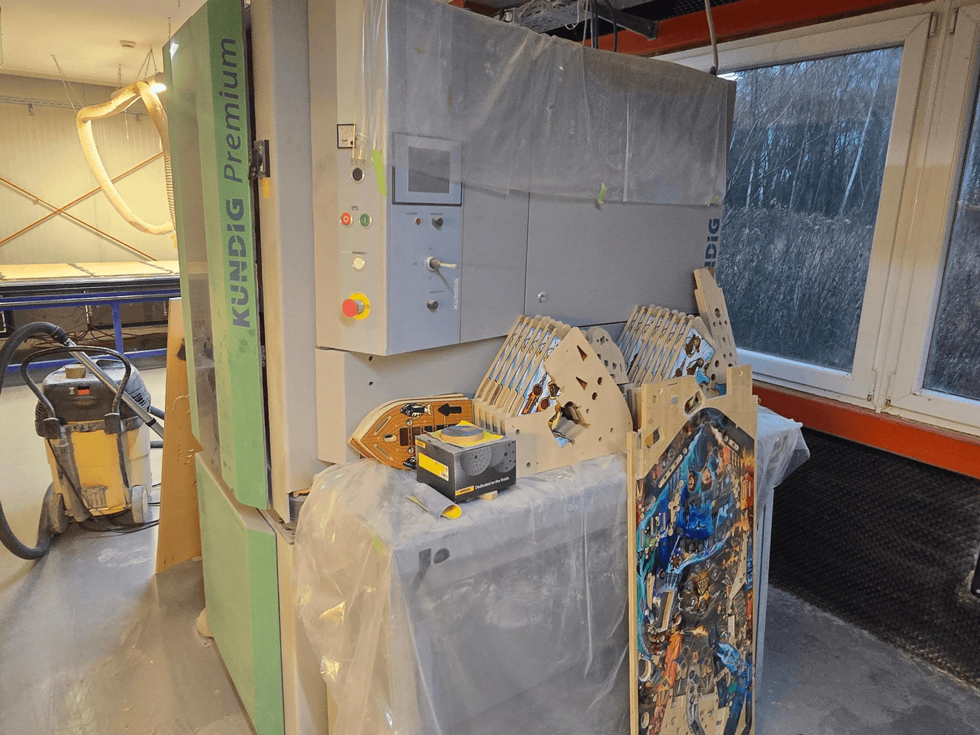 Szlifierka taśmowa Kundig Premium-1 1100-C w warsztacie, pokazująca panel sterowania i elementy drewniane, widok z boku, z odkurzaczem w tle.