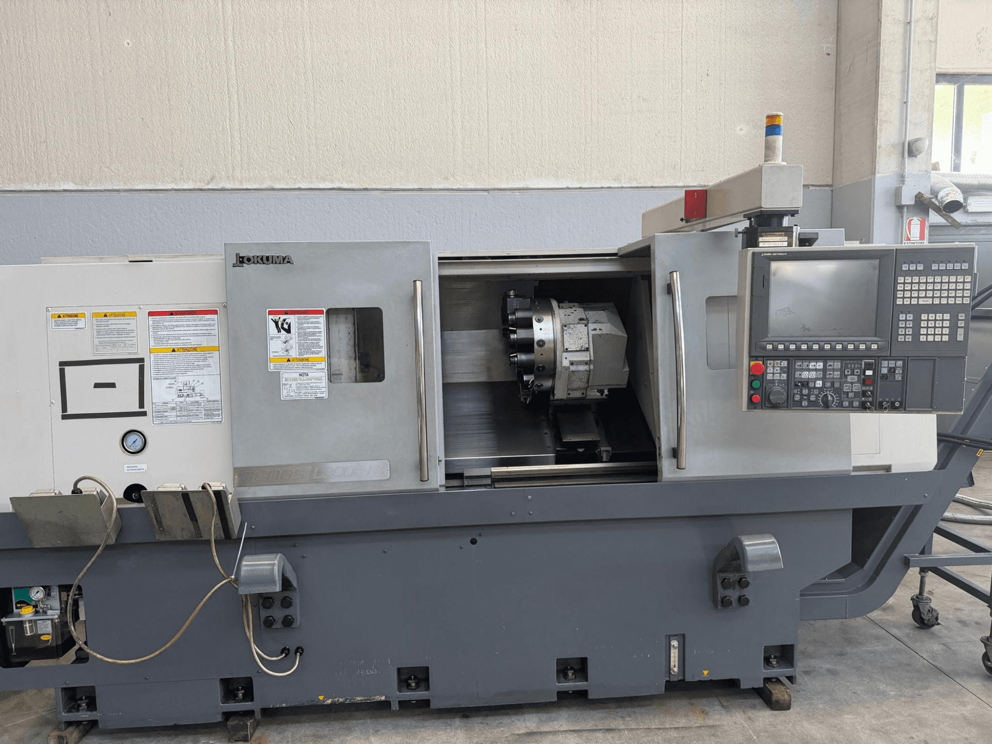 Tokarka CNC Okuma, widok z przodu, pokazujący panel sterowania, uchwyt narzędzia tnącego i ostrzeżenia dotyczące bezpieczeństwa z boku.