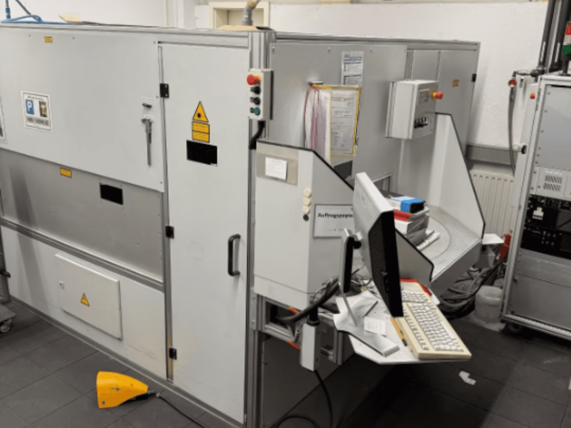 Maszyna przemysłowa z terminalem komputerowym, widok z boku; zawiera przyciski sterujące i znaki ostrzegawcze bezpieczeństwa.