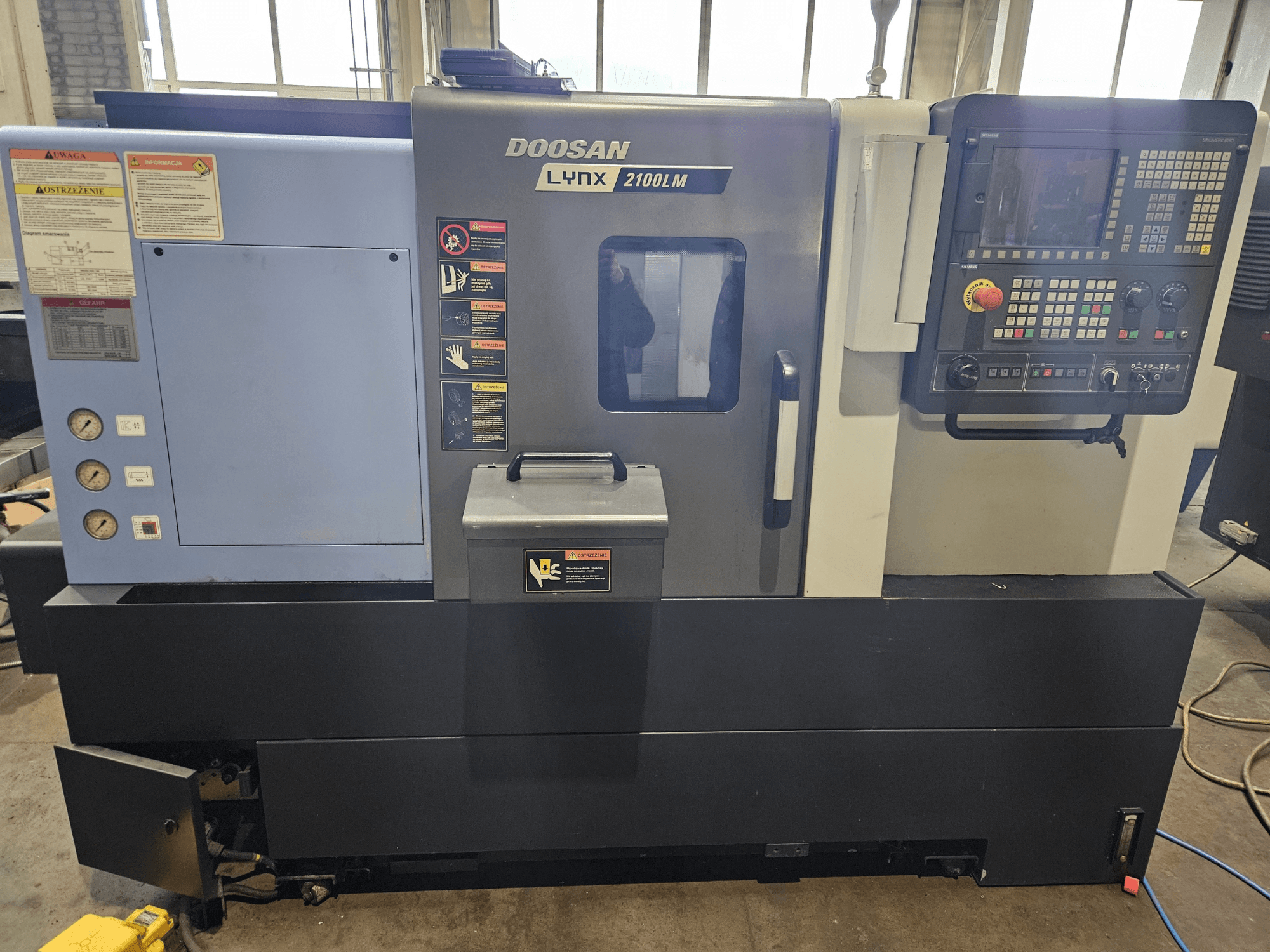 Tokarka CNC Doosan Lynx 2100LM, widok z przodu, widoczny panel sterowania, wskaźniki ciśnienia powietrza i etykiety bezpieczeństwa.