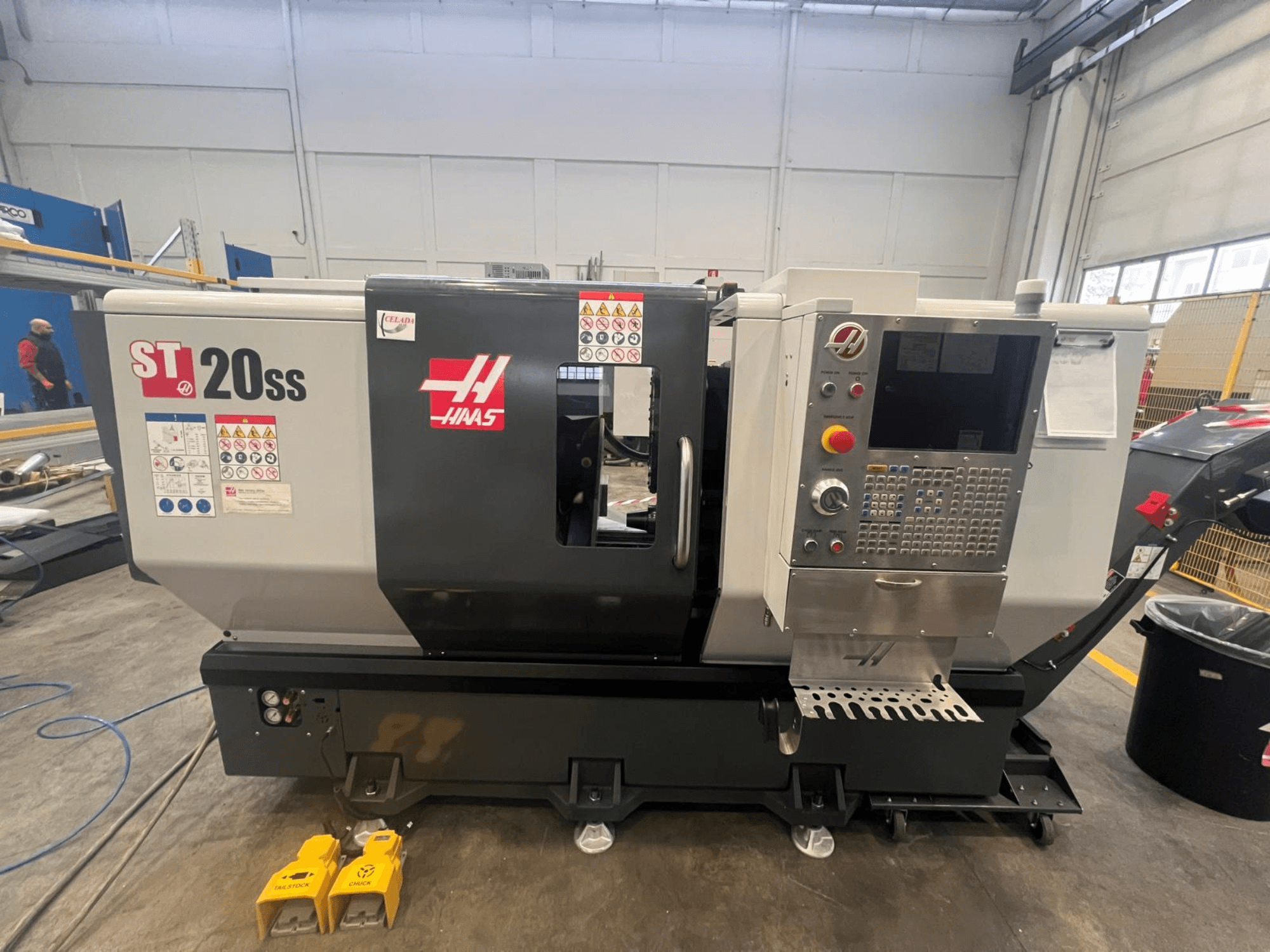 Tokarka CNC Haas ST 20SS w widoku z boku, z panelem sterowania, etykietami bezpieczeństwa i pedałami na podłodze.