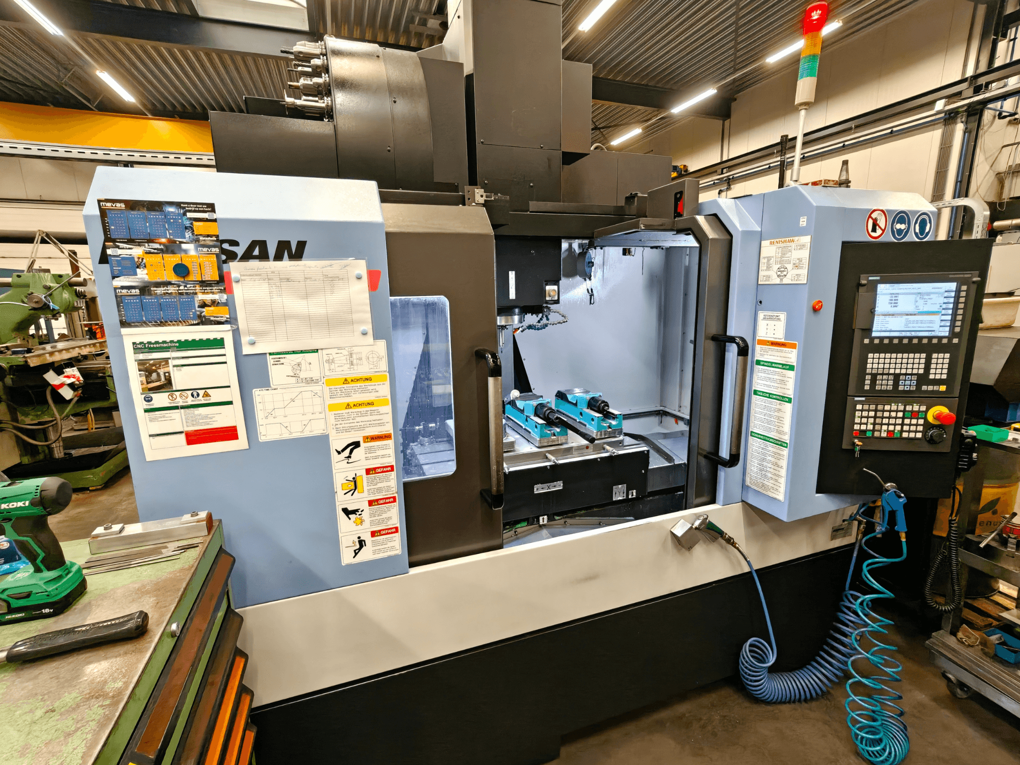 Pionowe centrum obróbcze DOOSAN DNM 400 II w warsztacie, z elementami sterującymi, uchwytami narzędziowymi i oznakowaniem bezpieczeństwa, widok z przodu.
